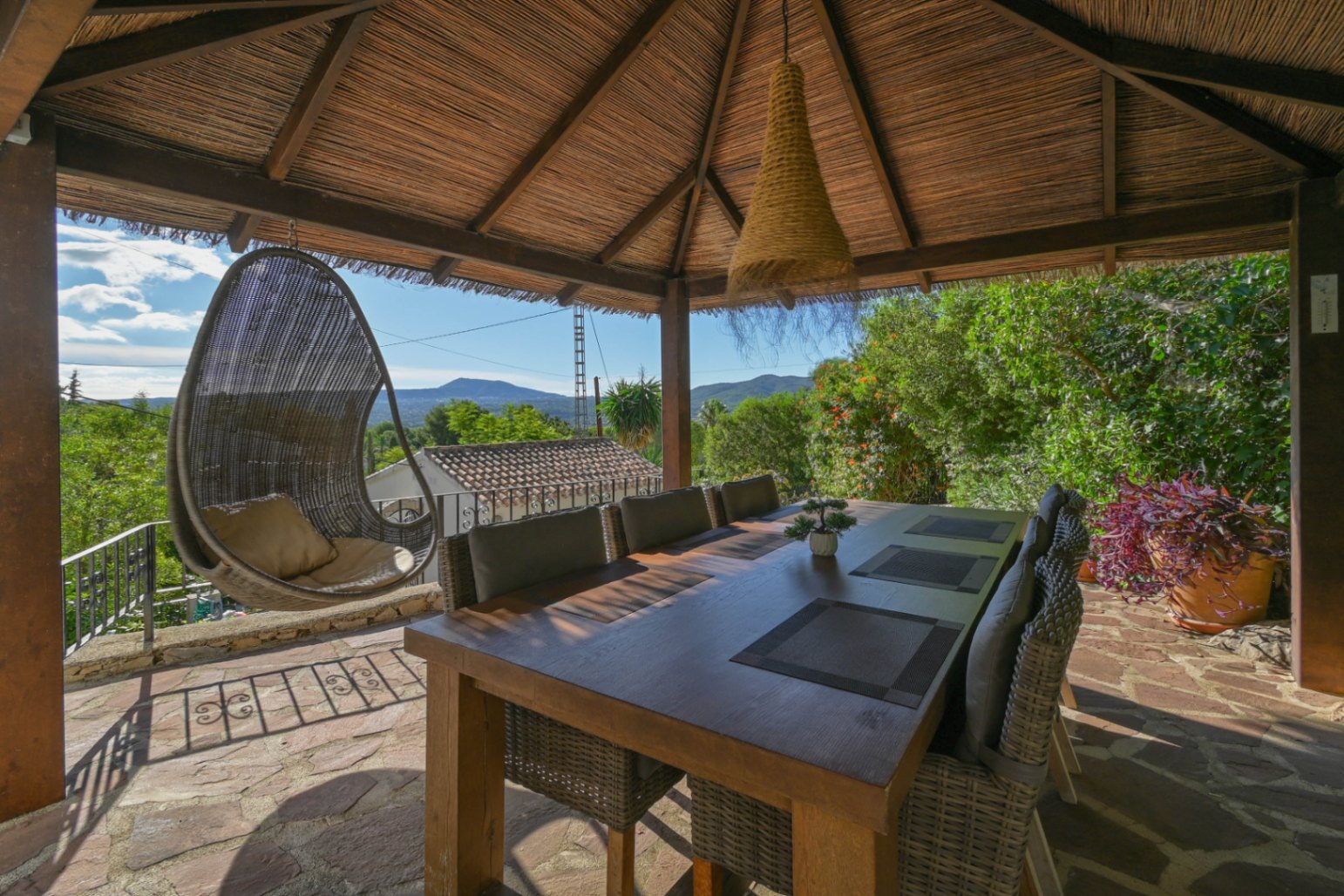 Außenessbereich unter einem Reetdach-Pavillon mit Holztisch, Korbstühlen und einem Hängesessel mit Blick auf die umliegende Berglandschaft.
