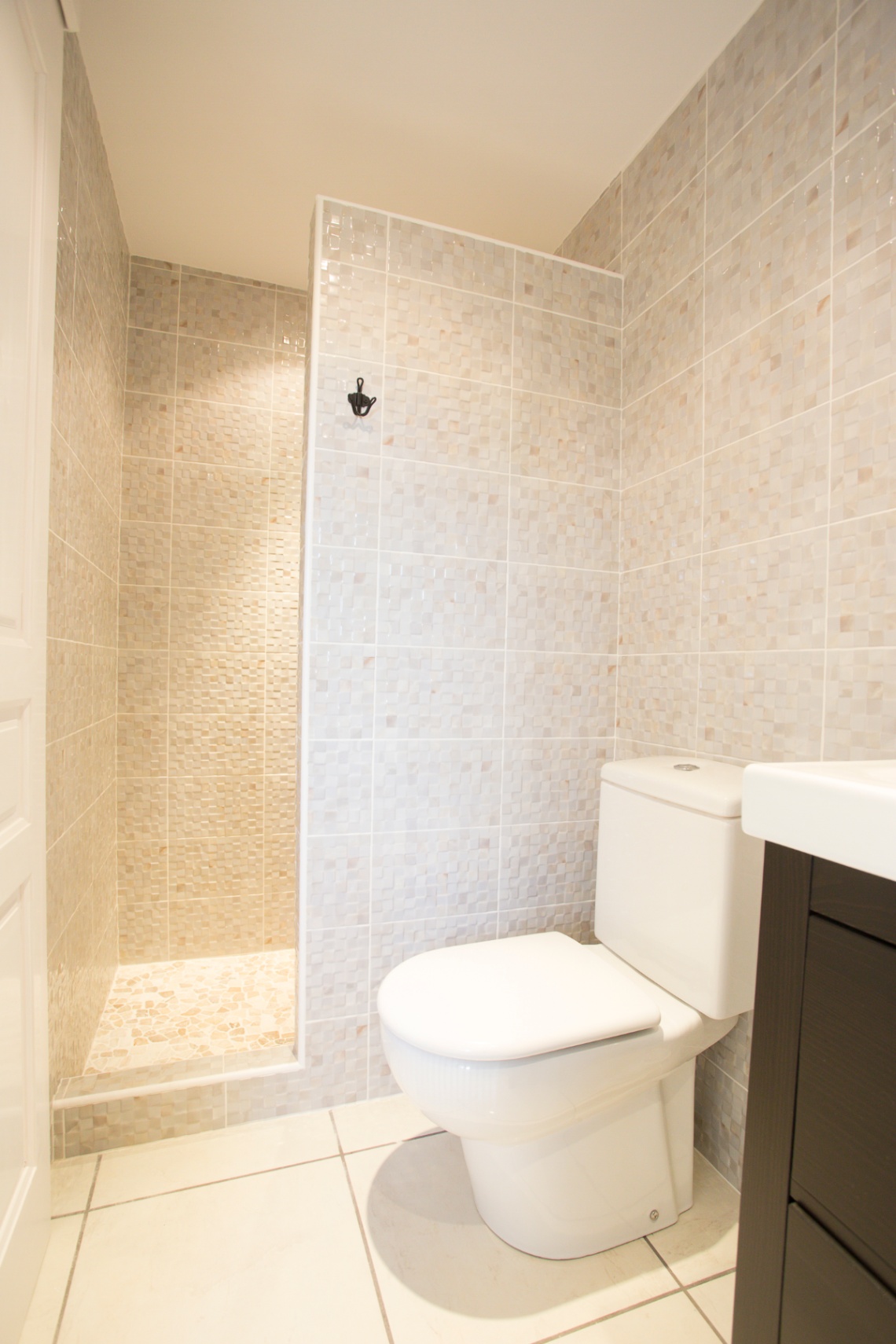 Badezimmer mit begehbarer Dusche mit beigen Mosaikfliesen, weißem WC und dunklem Waschtischunterschrank.