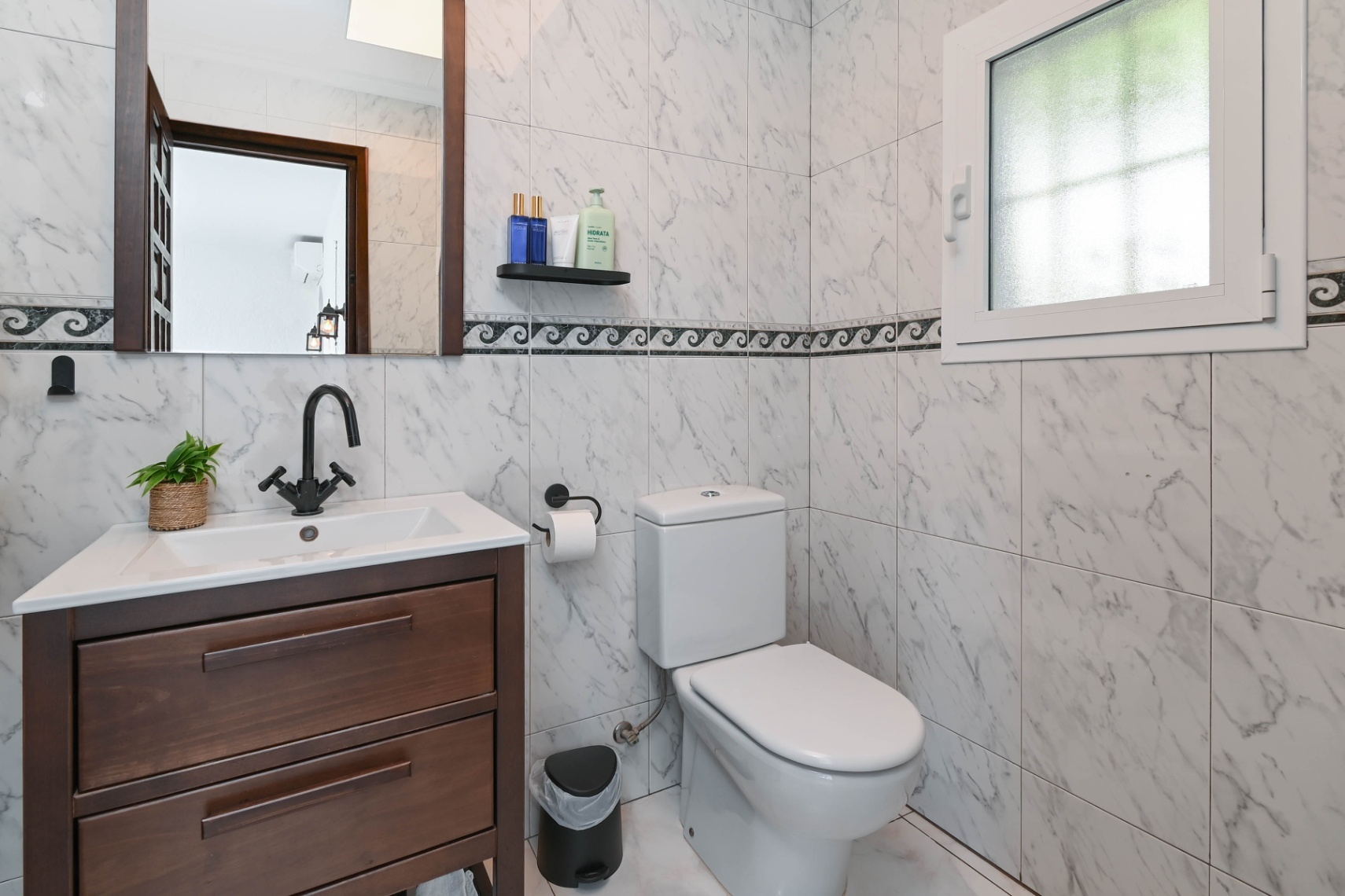 Badezimmer mit marmorierten Wandfliesen, dunklem Holzwaschtisch, mattschwarzen Armaturen und einem kleinen Milchglasfenster für optimale Privatsphäre.