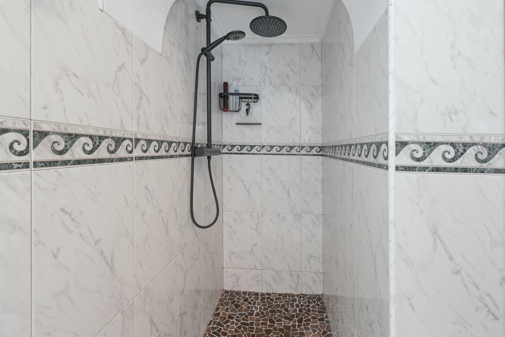 Begehbare Dusche mit Wandfliesen in Marmoroptik, Wellendekor-Bordüre, Kieselsteinboden und einem modernen Duschsystem in Mattschwarz.