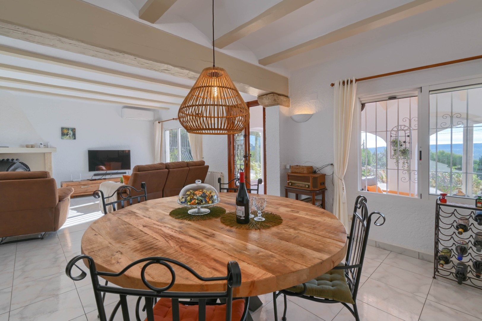Essbereich mit rundem Holztisch, schmiedeeisernen Stühlen und Korbpendelleuchte unter einer Decke mit sichtbaren Holzbalken und Blick ins Wohnzimmer.