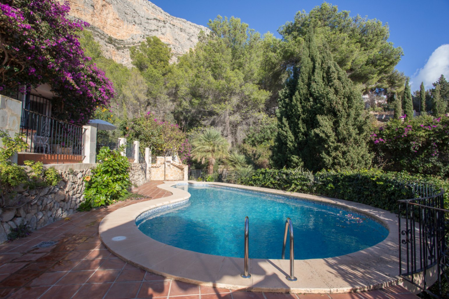 Geschwungener Pool mit blauen Mosaikfliesen, umgeben von Terrakotta-Terrassen und mediterraner Bepflanzung vor einer markanten Felswand.