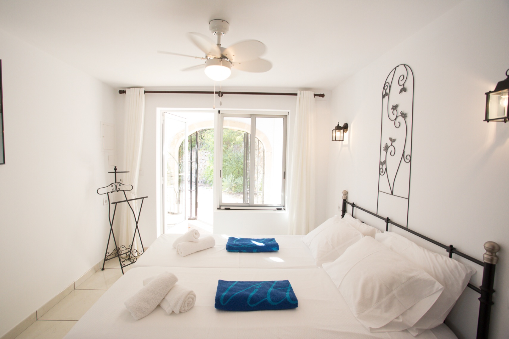 Helles Schlafzimmer mit weißen Wänden, schmiedeeisernem Bettgestell und Deckenventilator. Glastüren bieten direkten Zugang zu einer sonnigen Terrasse.