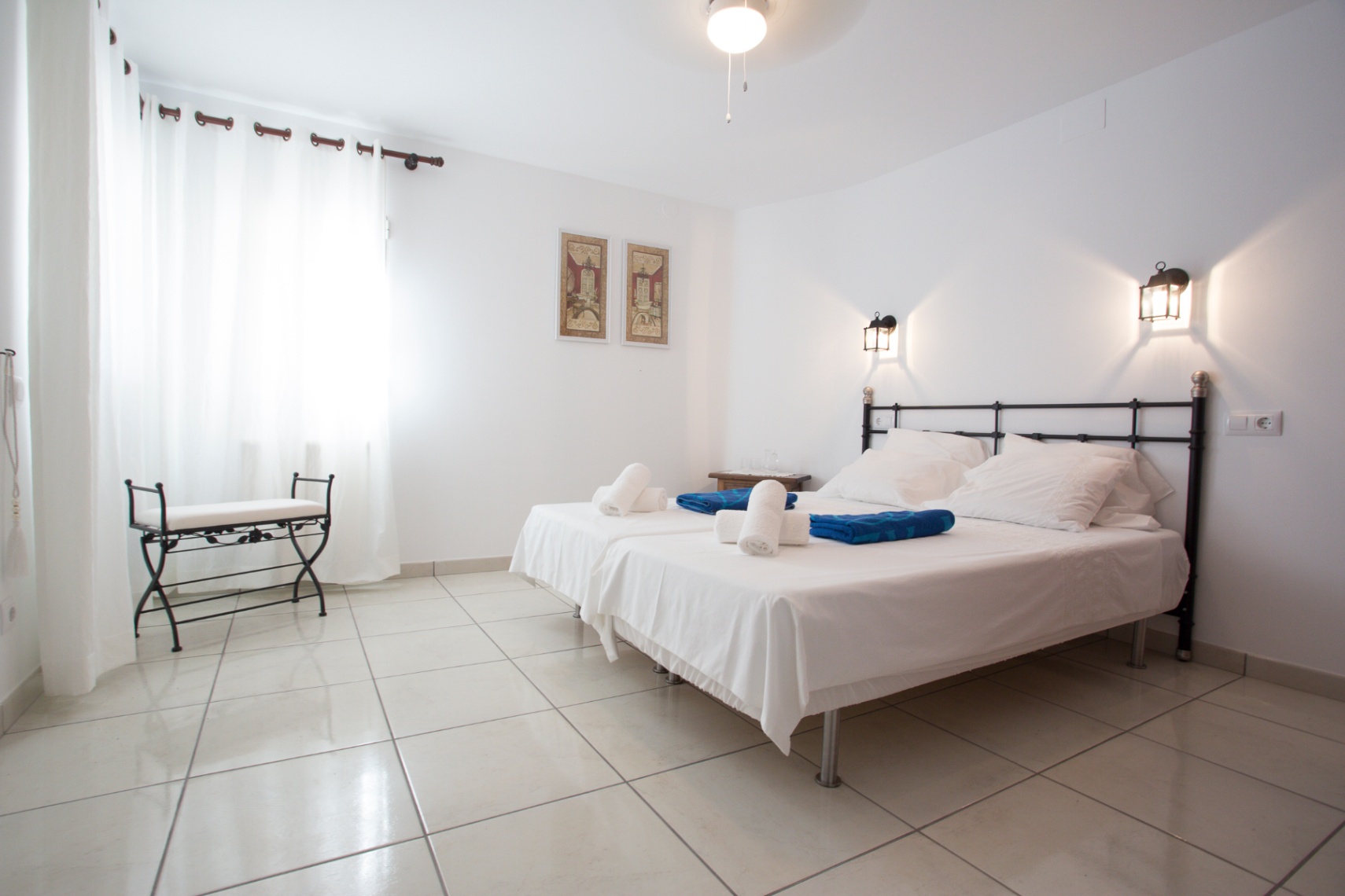 Helles Schlafzimmer mit zwei Einzelbetten, schmiedeeisernem Kopfteil und Fliesenboden. Wandlaternen und eine passende Eisenbank ergänzen das Design.