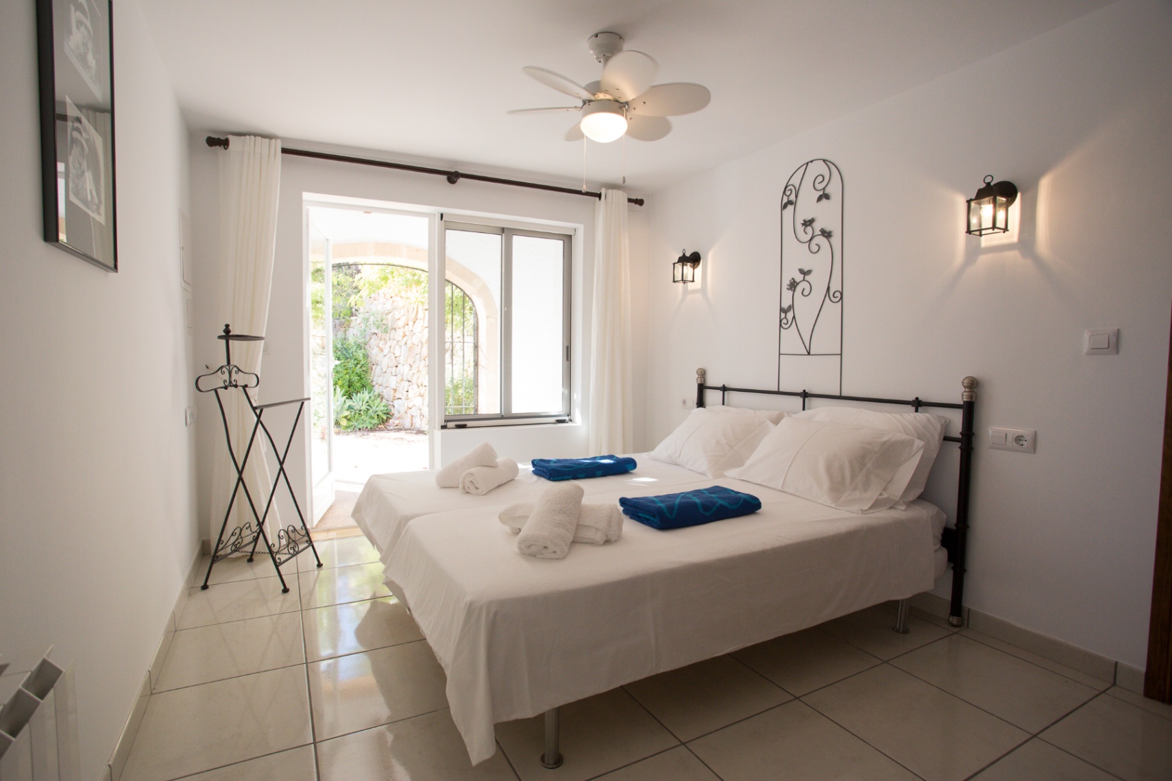 Helles Schlafzimmer mit zwei Einzelbetten, schmiedeeisernen Details und Deckenventilator. Die Glastür bietet direkten Zugang zu einem privaten Innenhof.