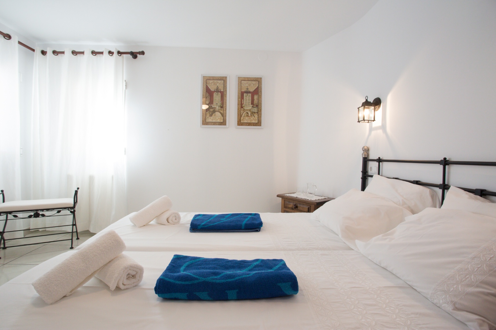 Helles Schlafzimmer mit zwei Einzelbetten, schwarzem Schmiedeeisen-Kopfteil und passender Bank. Ein Nachttisch aus Holz und natürliches Licht ergänzen den Raum.