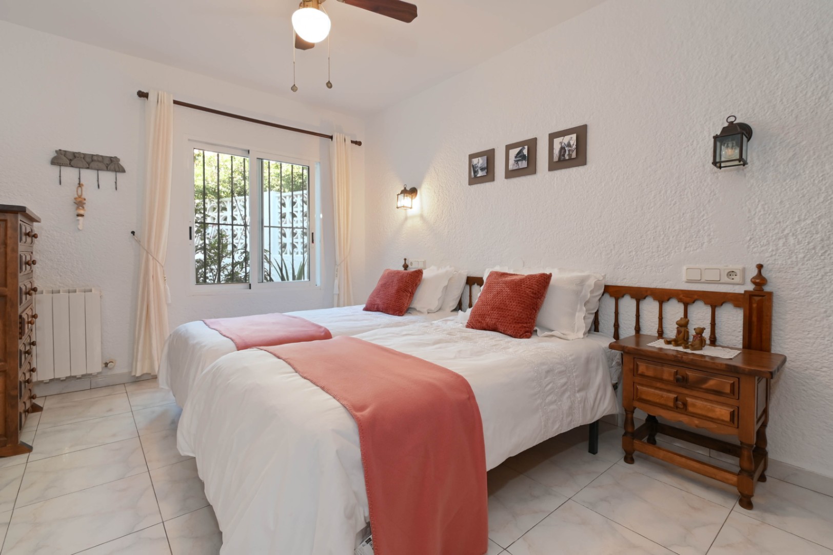 Helles Schlafzimmer mit zwei Einzelbetten, terrakottafarbenen Akzenten und rustikalen Holzmöbeln. Strukturierte weiße Wände und ein Fenster mit Blick ins Grüne prägen den Raum.