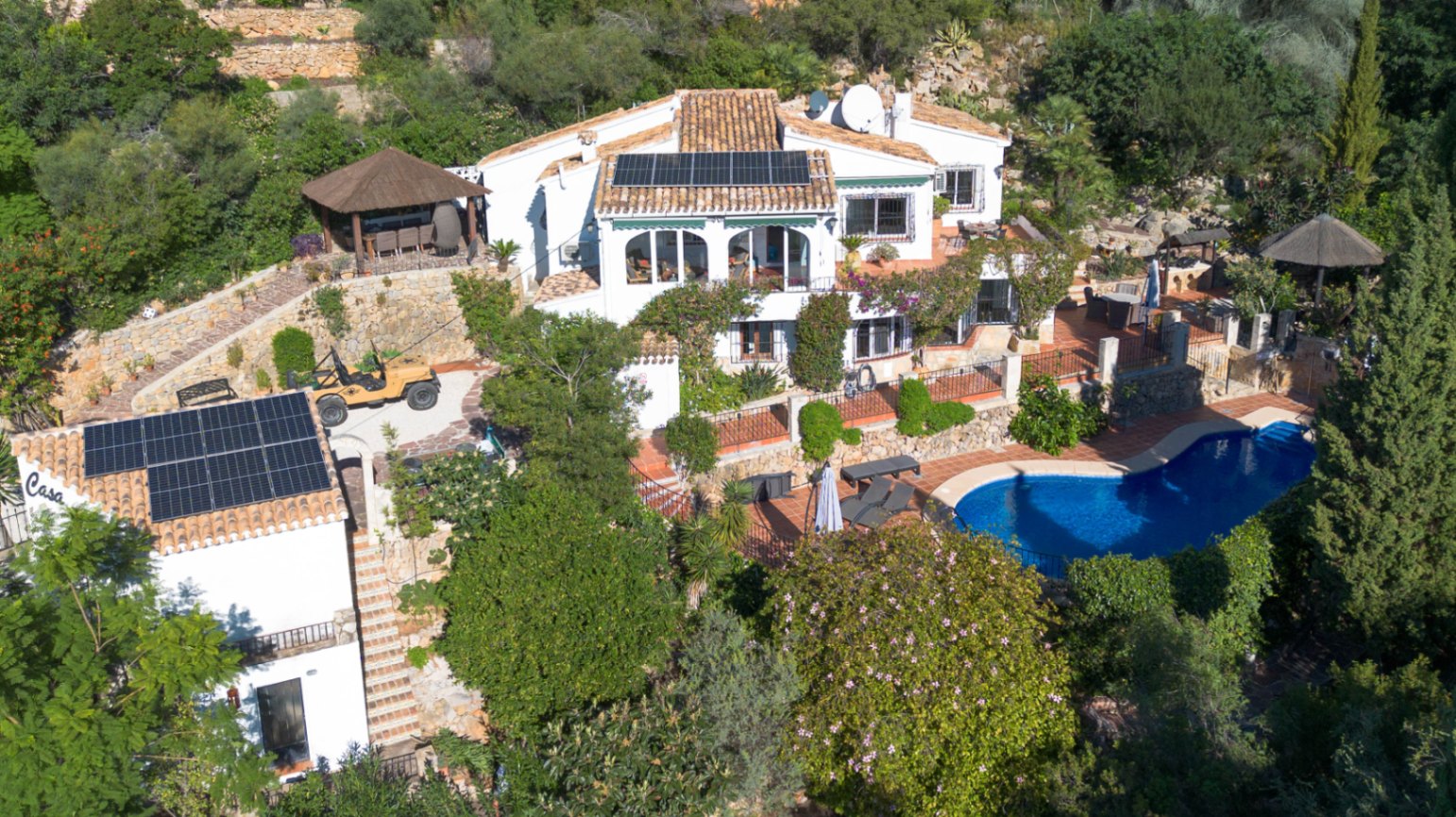 Luftaufnahme einer mediterranen Villa mit Solarmodulen auf Terrakottadächern, nierenförmigem Pool und terrassierten Gärten mit Natursteinmauern.