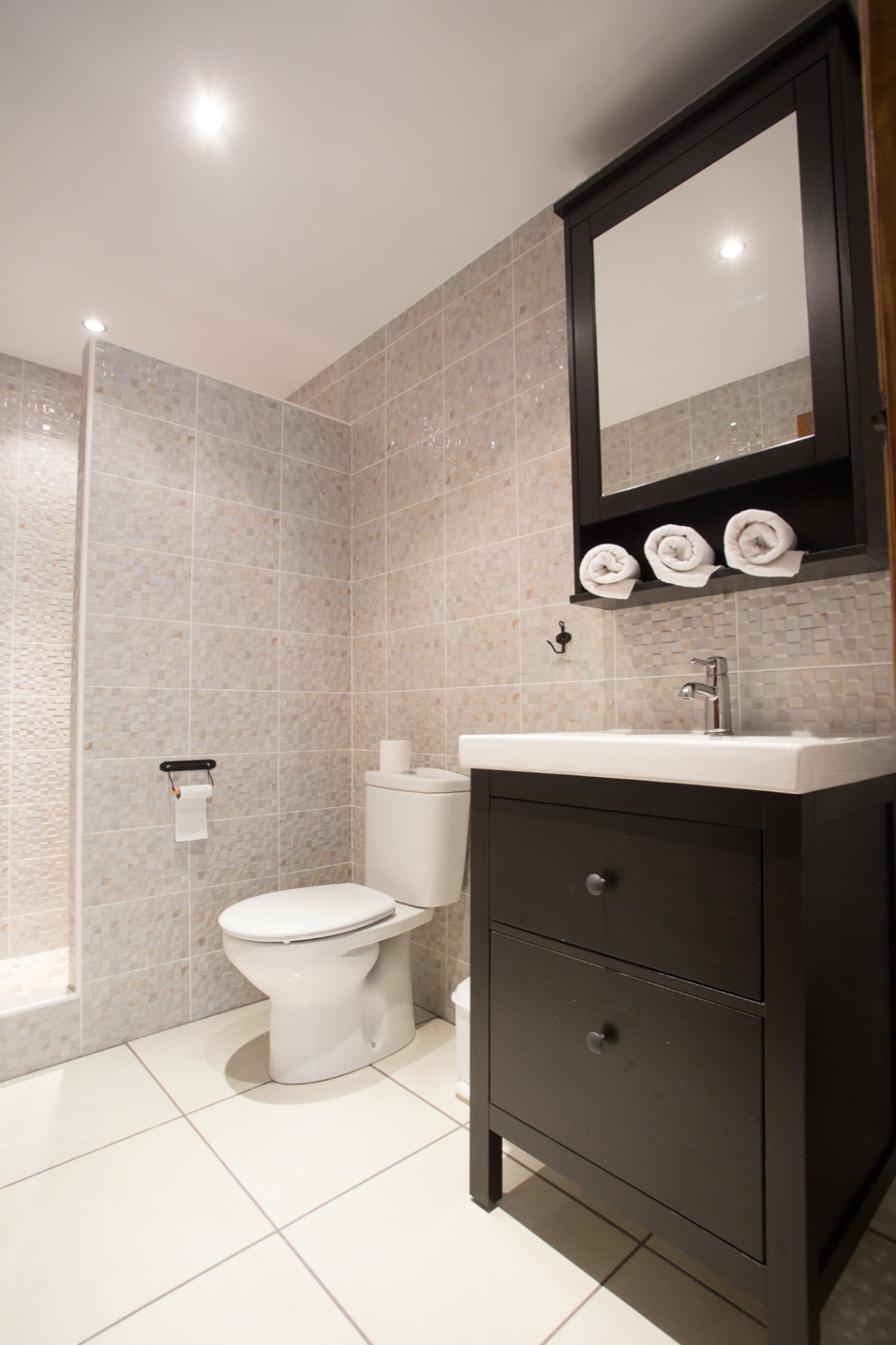 Modernes Badezimmer mit beigen Mosaikfliesen, einem dunklen Waschtisch mit weißem Keramikbecken und passendem Spiegelschrank mit Handtuchablage.