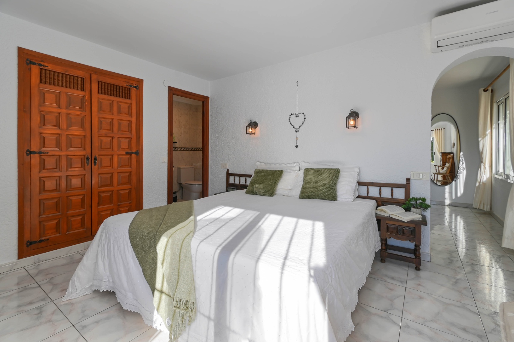 Schlafzimmer mit geschnitztem Holzeinbauschrank, hellem Marmorboden und En-Suite-Bad. Natürliches Licht beleuchtet das Bett mit grünen Akzenten und Wandleuchten im Laternenstil.