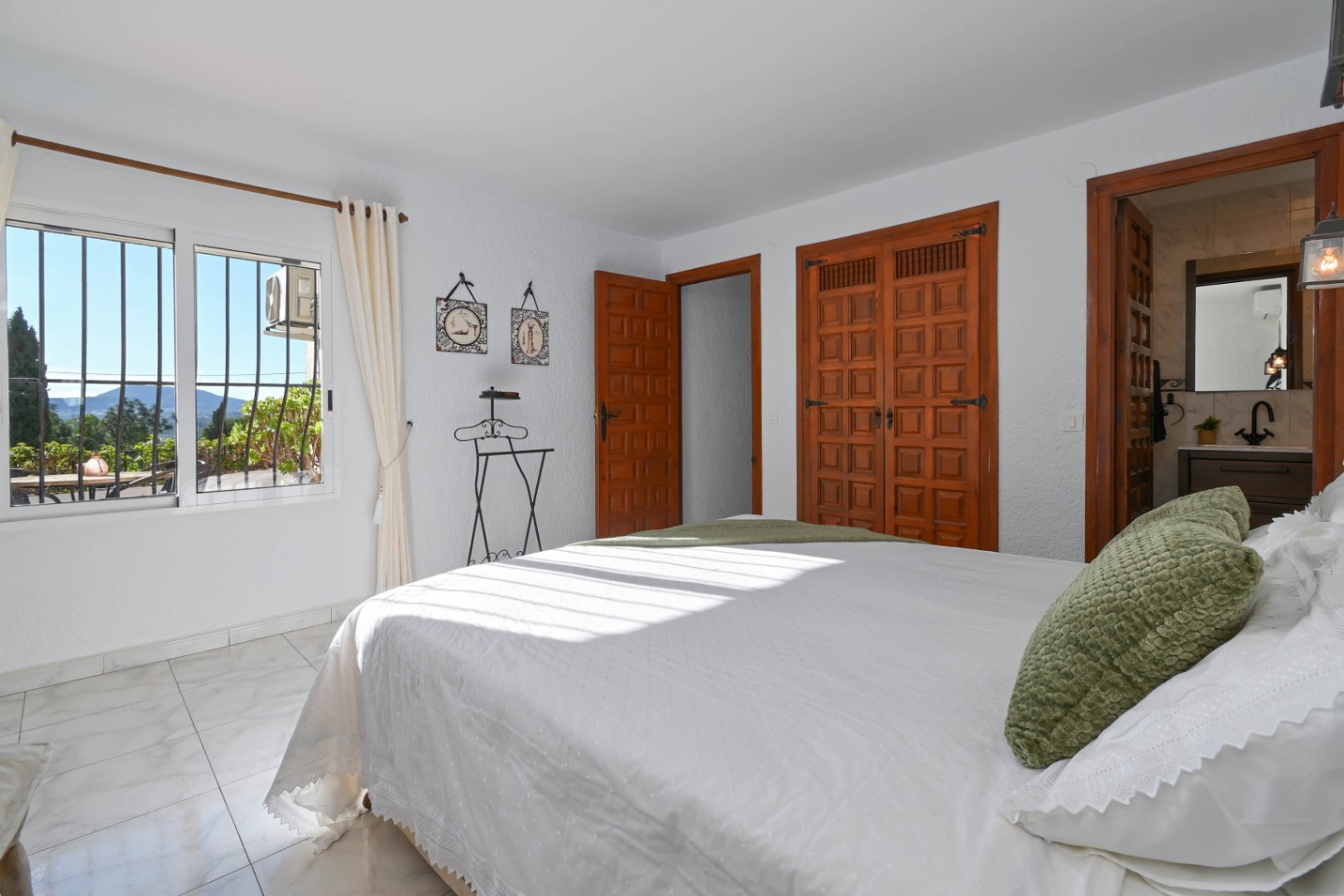 Schlafzimmer mit traditionellen Einbauschränken aus Holz, Marmoreffekt-Fliesenboden und angrenzendem Badezimmer. Das Fenster bietet einen Blick auf die Landschaft.