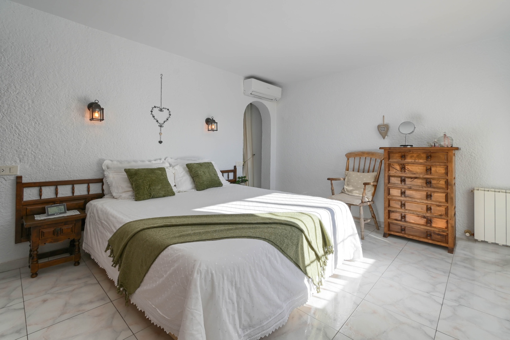 Schlafzimmer mit weiß verputzten Wänden, Marmorboden und rustikalen Holzmöbeln. Wandlaternen und grüne Akzente ergänzen das helle Ambiente.
