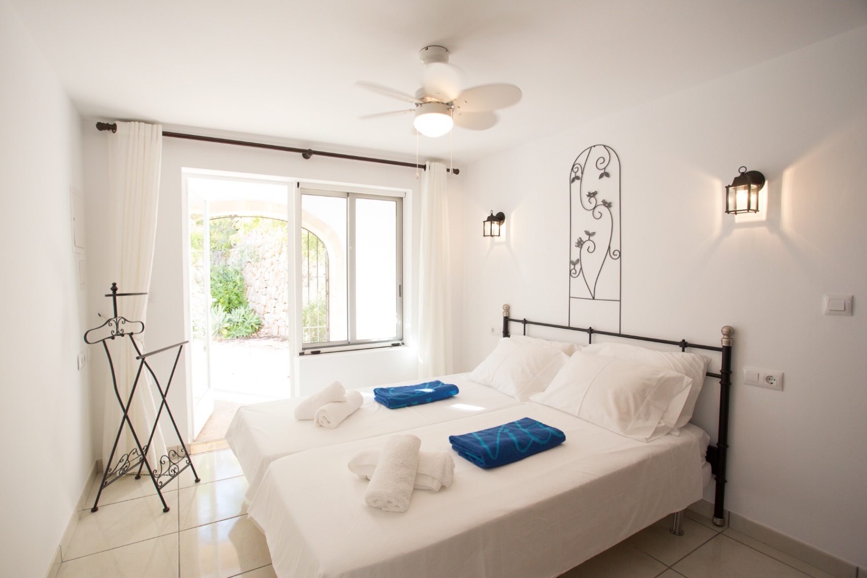 Schlafzimmer mit zwei Einzelbetten, schmiedeeisernem Kopfteil und Wandlaternen. Mit Deckenventilator und direktem Zugang zu einer Terrasse mit Steinmauern.