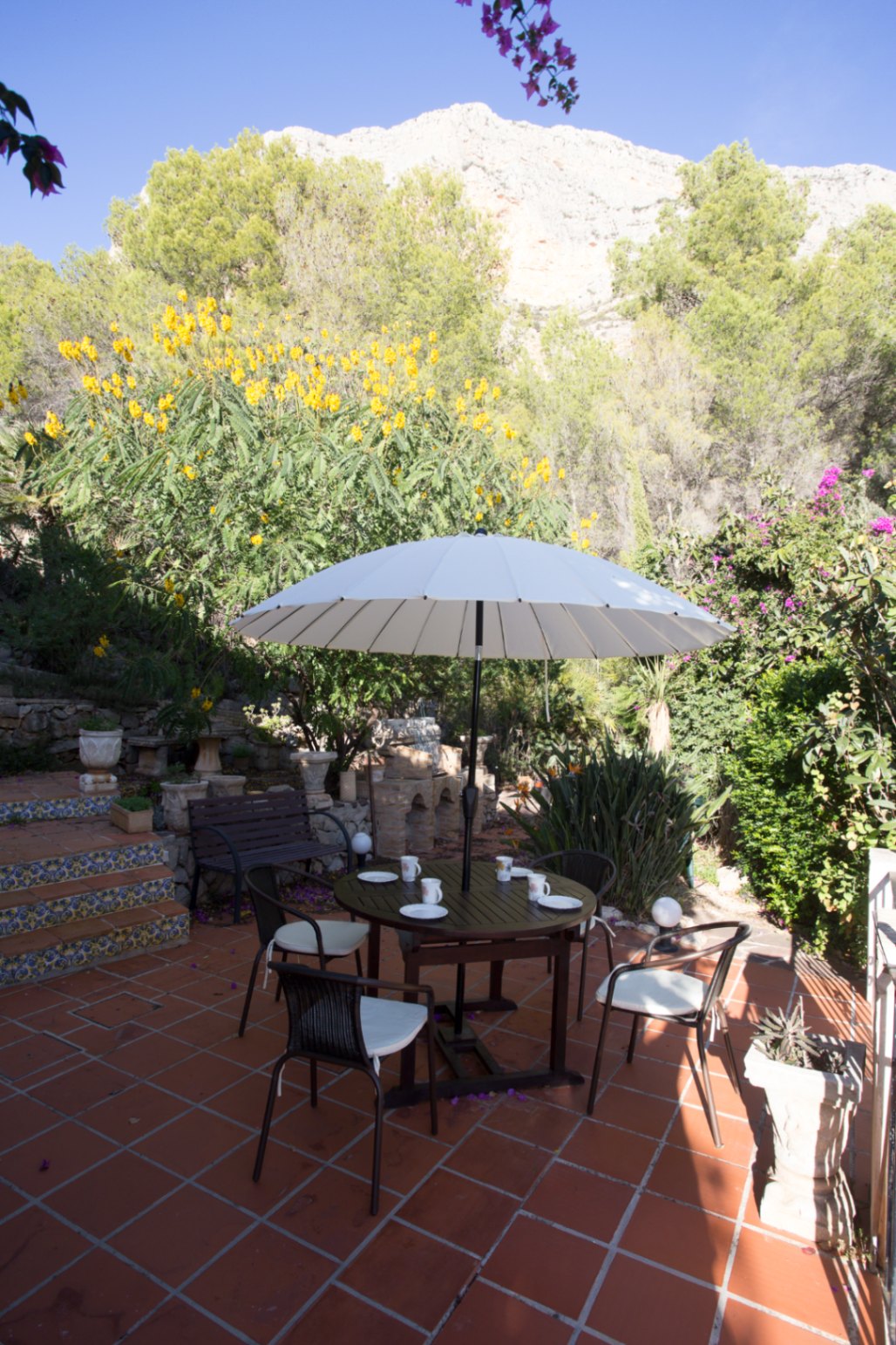 Terrakotta-Terrasse mit rundem Esstisch, weißem Sonnenschirm und Blick auf einen mediterranen Garten vor einer markanten Felswand.