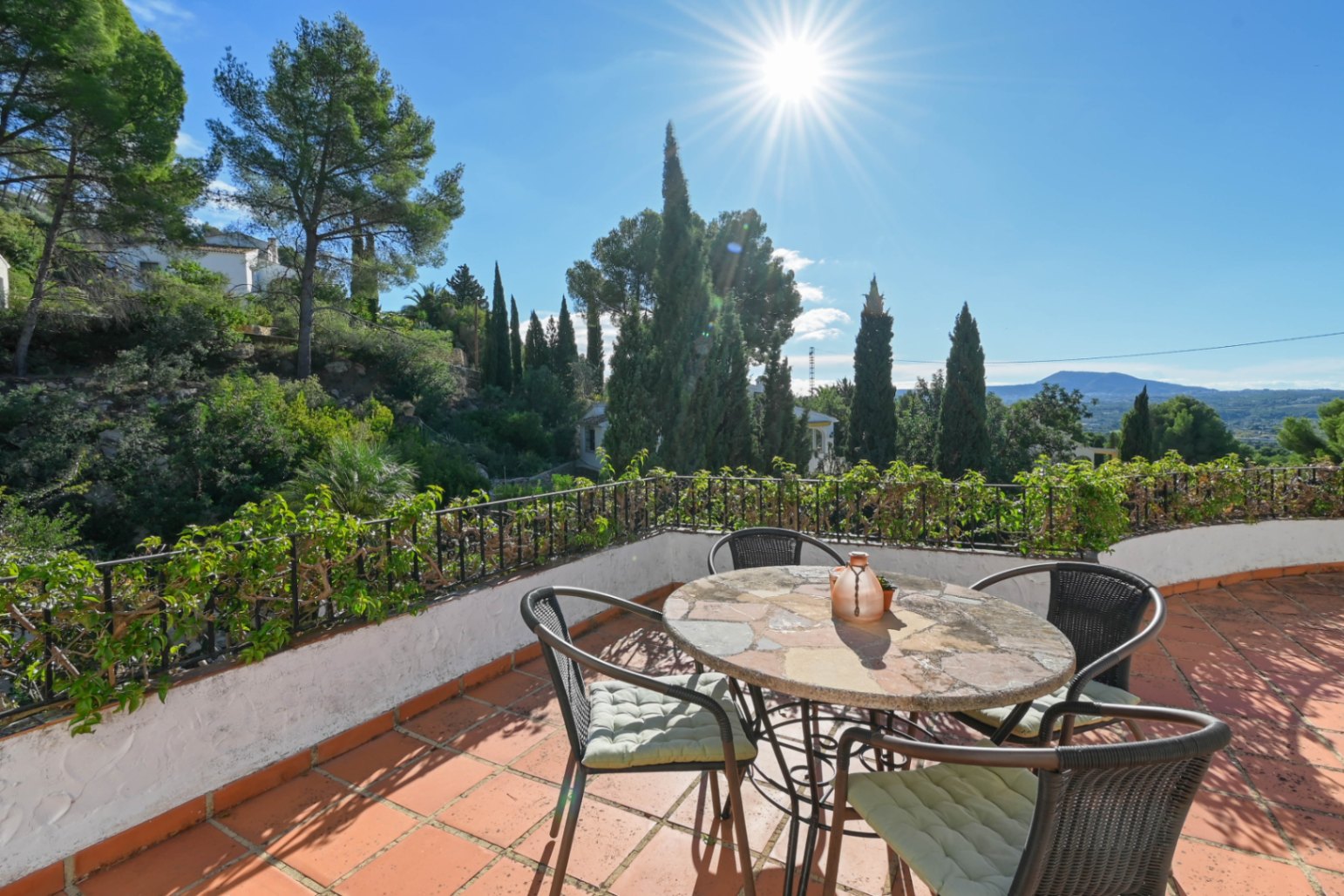 Terrasse mit Terrakottaboden, Steintisch und Korbstühlen, mit Blick auf mediterrane Vegetation und Berge unter strahlendem Sonnenschein.