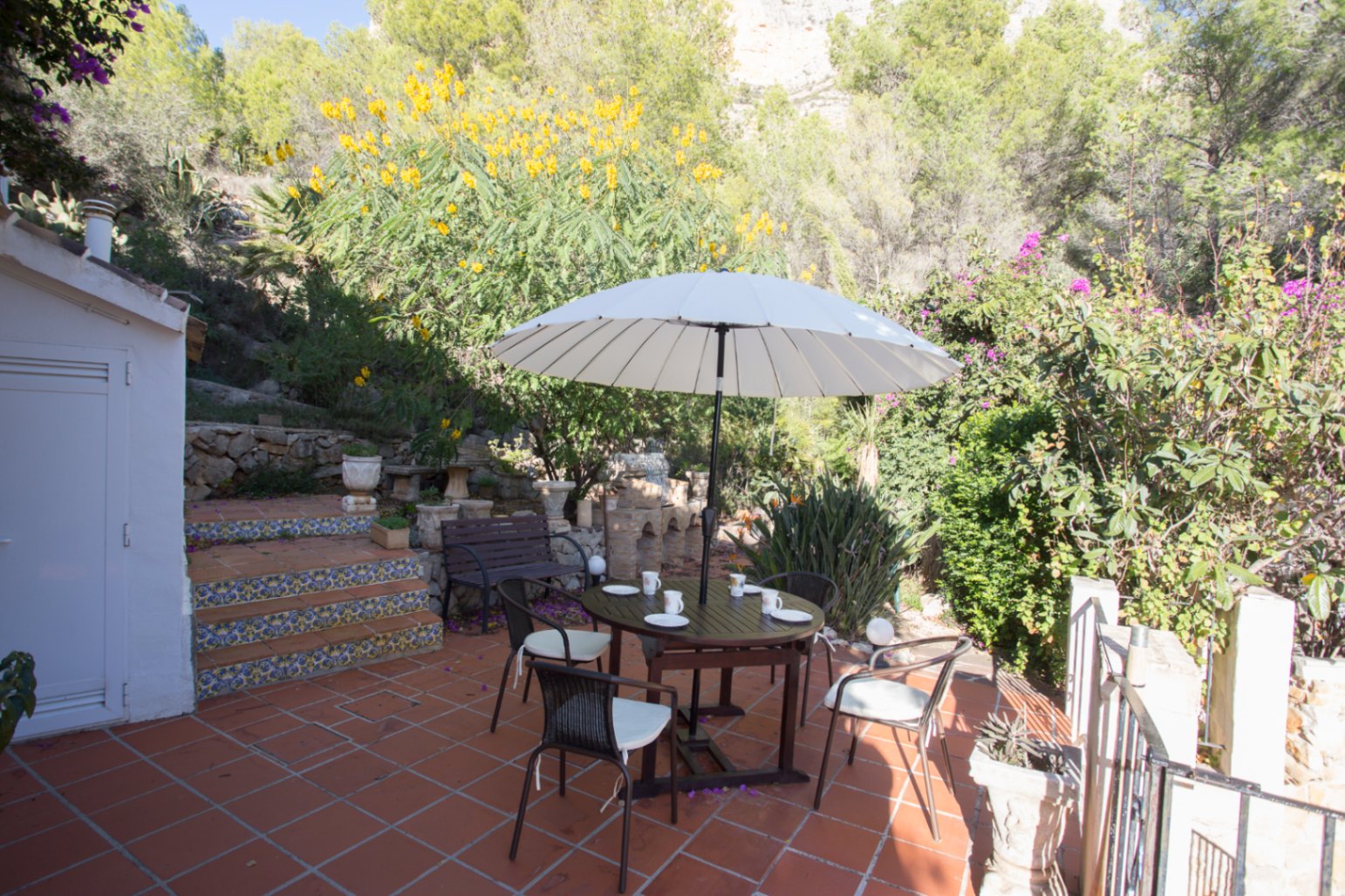 Terrasse mit Terrakottafliesen, Essbereich unter einem weißen Sonnenschirm und dekorativen Fliesentreppen vor einer bewaldeten Hügellandschaft.