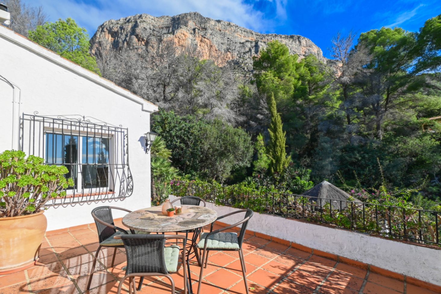 Terrasse mit Terrakottafliesen und Steintisch, mit Blick auf den angrenzenden Wald und die markante Felswand des Berges.