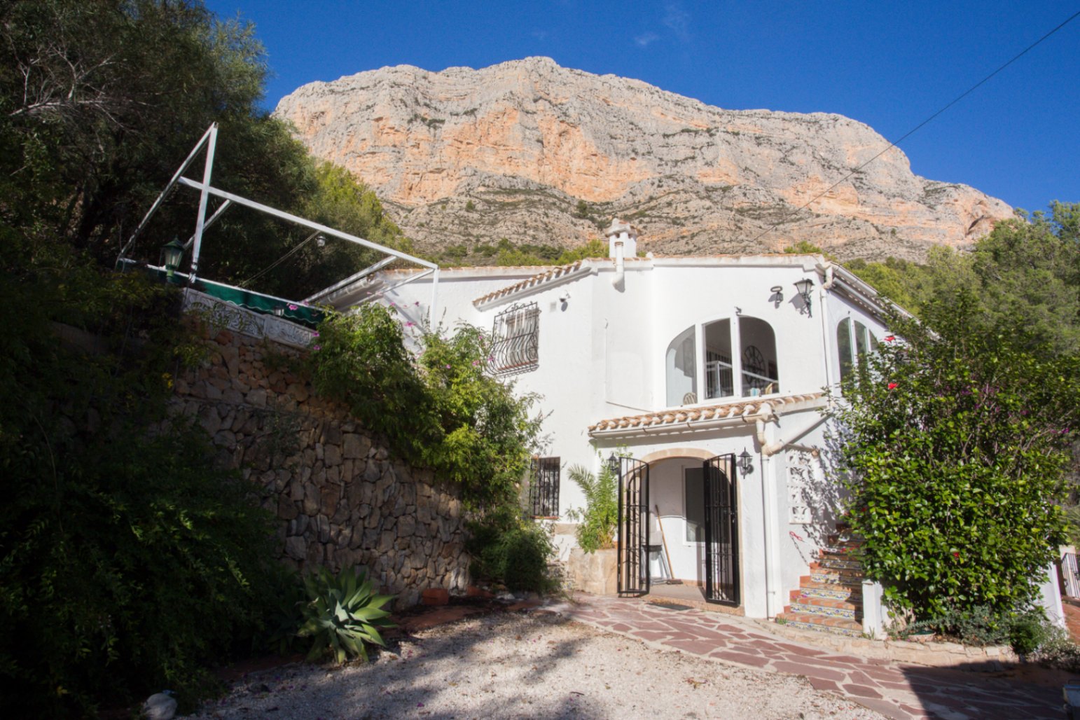 Weiße mediterrane Villa mit Rundbogenfenstern am Fuße eines massiven Felsmassivs, ergänzt durch Natursteinmauern und üppige Vegetation.