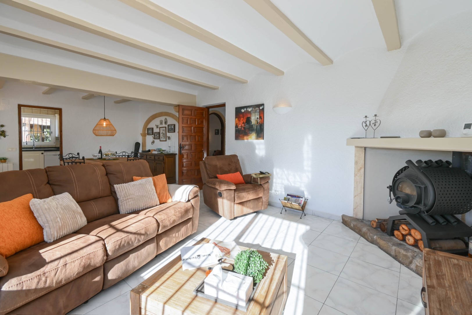 Wohnzimmer mit sichtbaren Deckenbalken, braunem Sofa und einem traditionellen Holzofen in einer Nische. Der Raum besticht durch helle Fliesen und natürliches Licht.