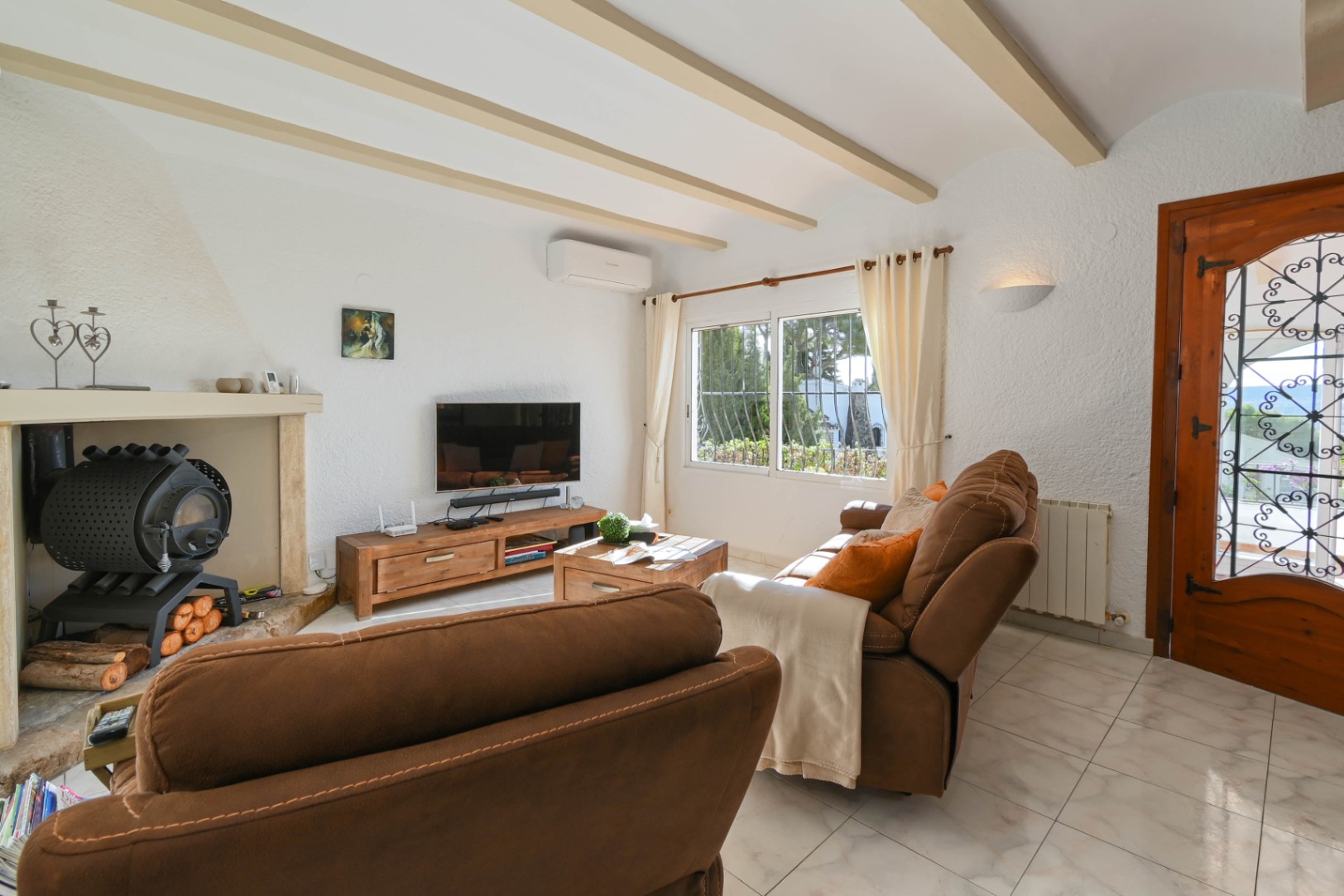 Wohnzimmer mit sichtbaren Deckenbalken, Kaminofen und rustikalen Holzmöbeln. Fliesenboden und Tageslicht durch das Fenster und die verzierte Glastür schaffen ein helles Ambiente.