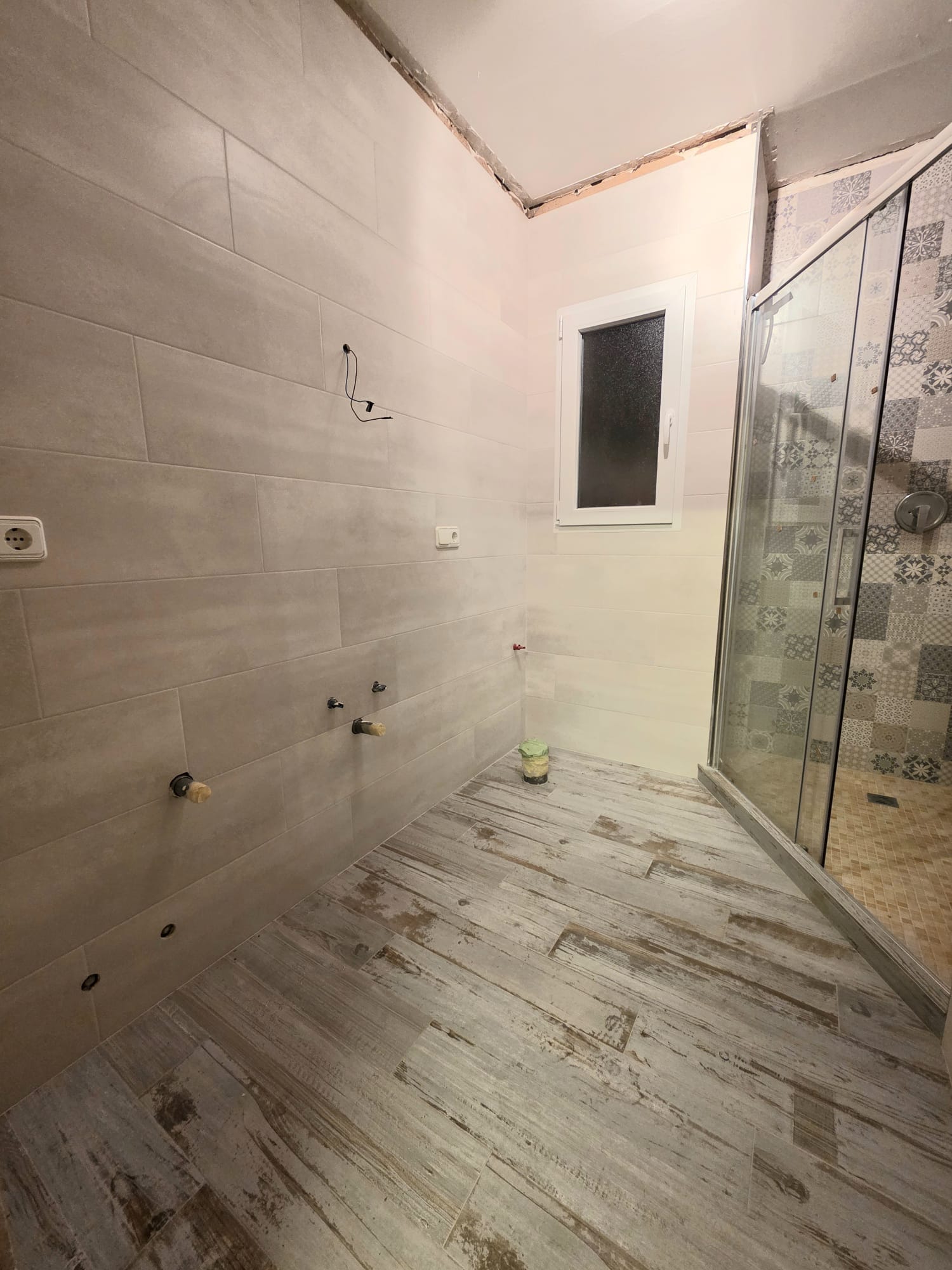 Badezimmer im Umbau mit Bodenfliesen in Holzoptik, großformatigen Wandfliesen und einer Glasduschkabine mit dekorativen Mustern im Innenbereich.