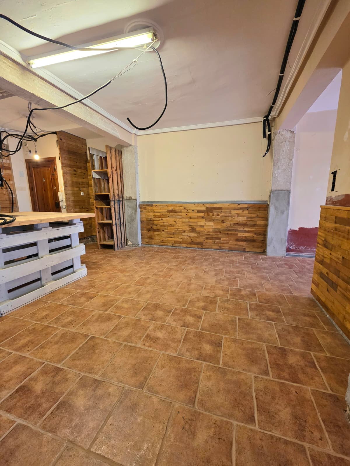 Innenraum mit Terrakotta-Fliesenboden, rustikaler Holzvertäfelung und einer Theke aus weiß gestrichenen Paletten mit heller Holzoberfläche.