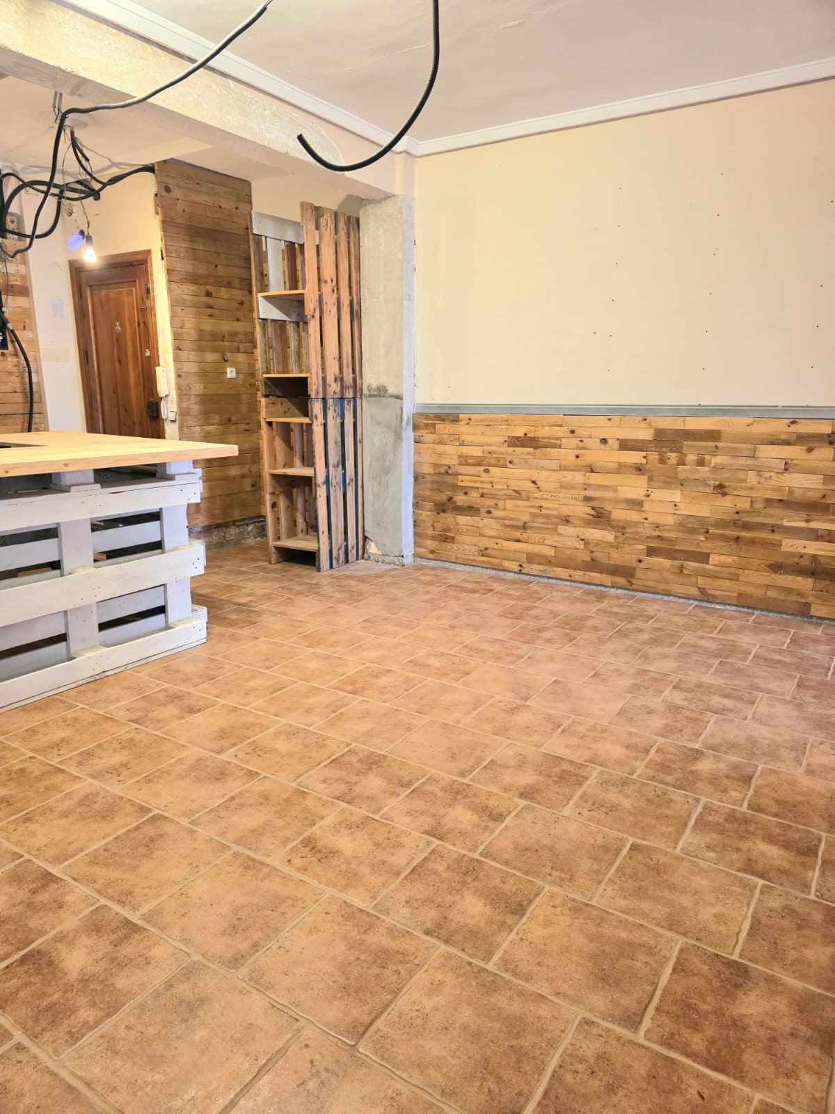 Innenraum mit Terrakotta-Fliesenboden und rustikaler Holzverkleidung. Ein Tresen aus weißen Paletten und Holzregale prägen den funktionalen Bereich.