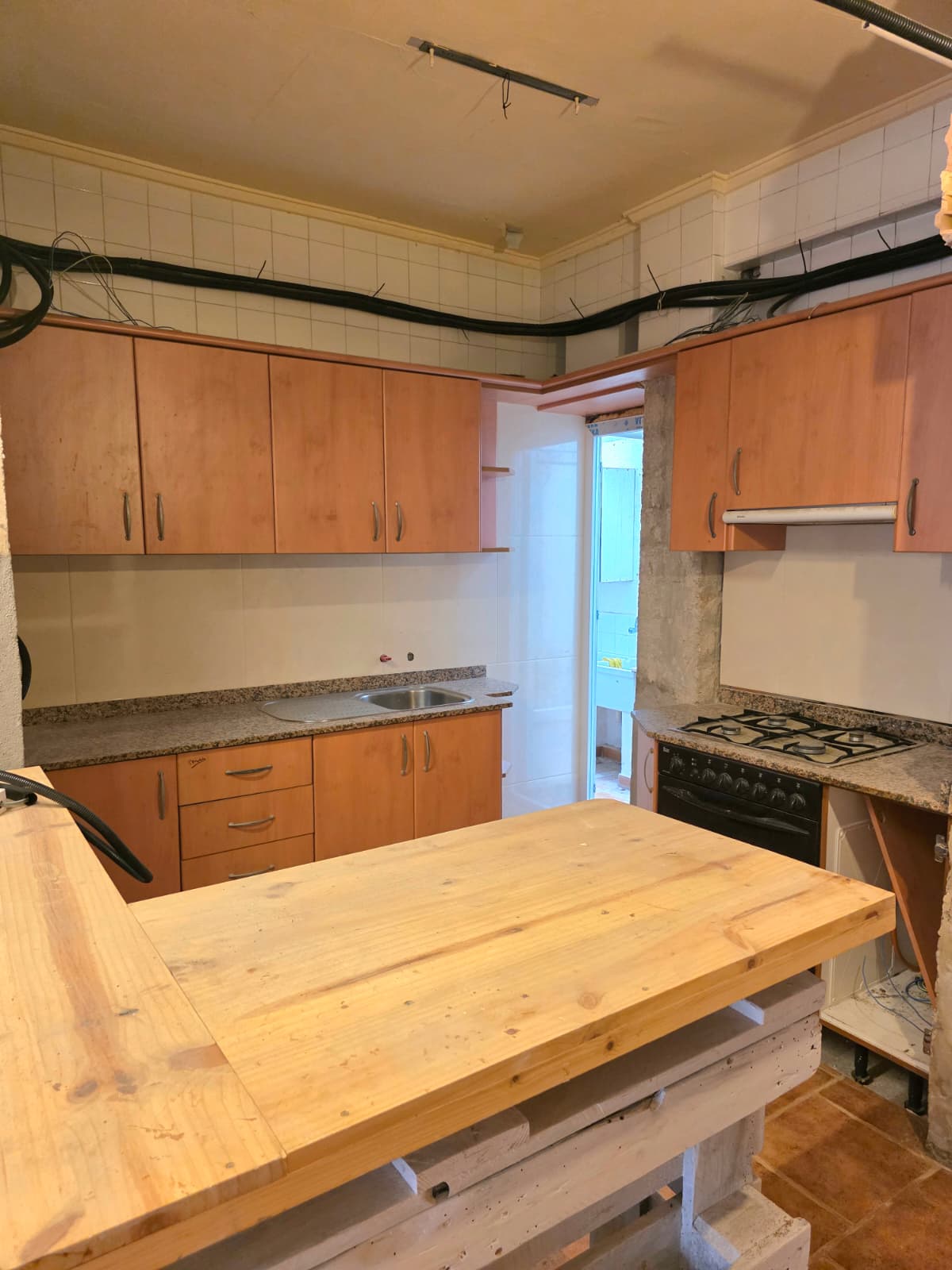 Küche mit Holzfronten, Granitarbeitsplatten und einer zentralen Holzinsel. Sichtbare Leitungen und geflieste Wände prägen den funktionalen Raum.