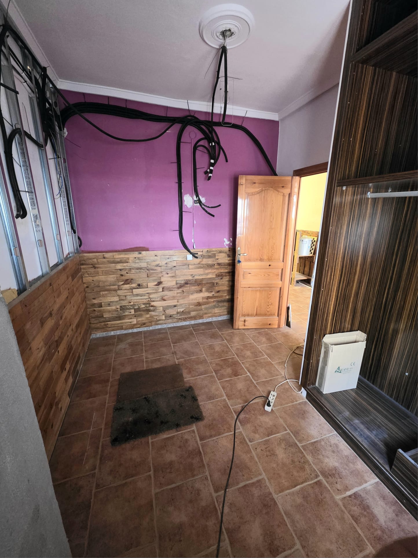 Renovierungsbedürftiger Raum mit Terrakotta-Fliesen, Holzverkleidung an den Wänden, lila Anstrich und offenliegenden Elektroinstallationen.