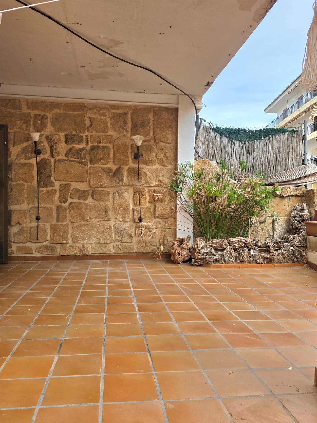 Überdachte Terrasse mit Terrakotta-Fliesen, einer rustikalen Natursteinwand mit Wandleuchten und einem dekorativen Pflanzbereich mit Felseinfassung.