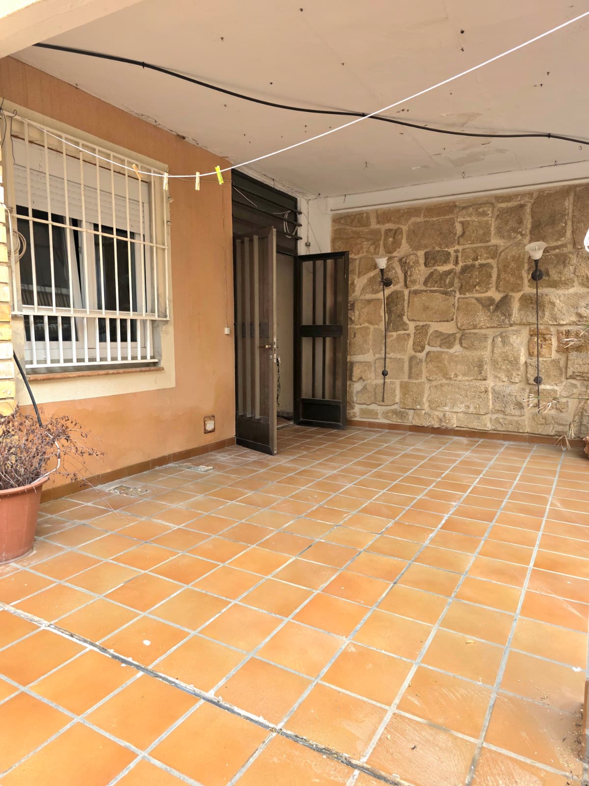 Überdachte Terrasse mit Terrakotta-Fliesenboden, einer rustikalen Natursteinwand mit Wandleuchten und einem weiß vergitterten Fenster.