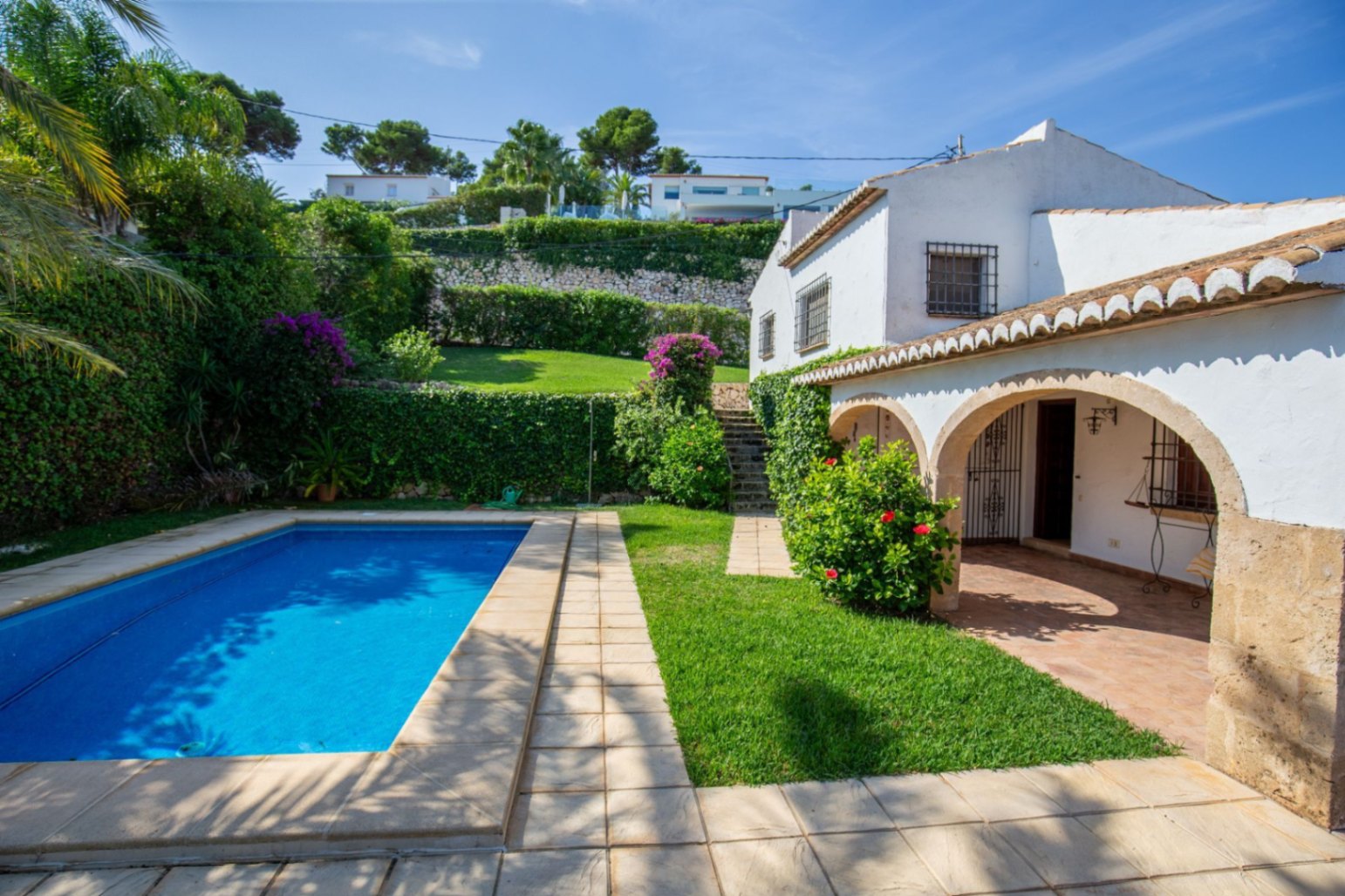Außenansicht einer mediterranen Villa mit rechteckigem Pool, steinernem Rundbogen-Portal und terrassiertem Garten mit üppiger Bepflanzung.