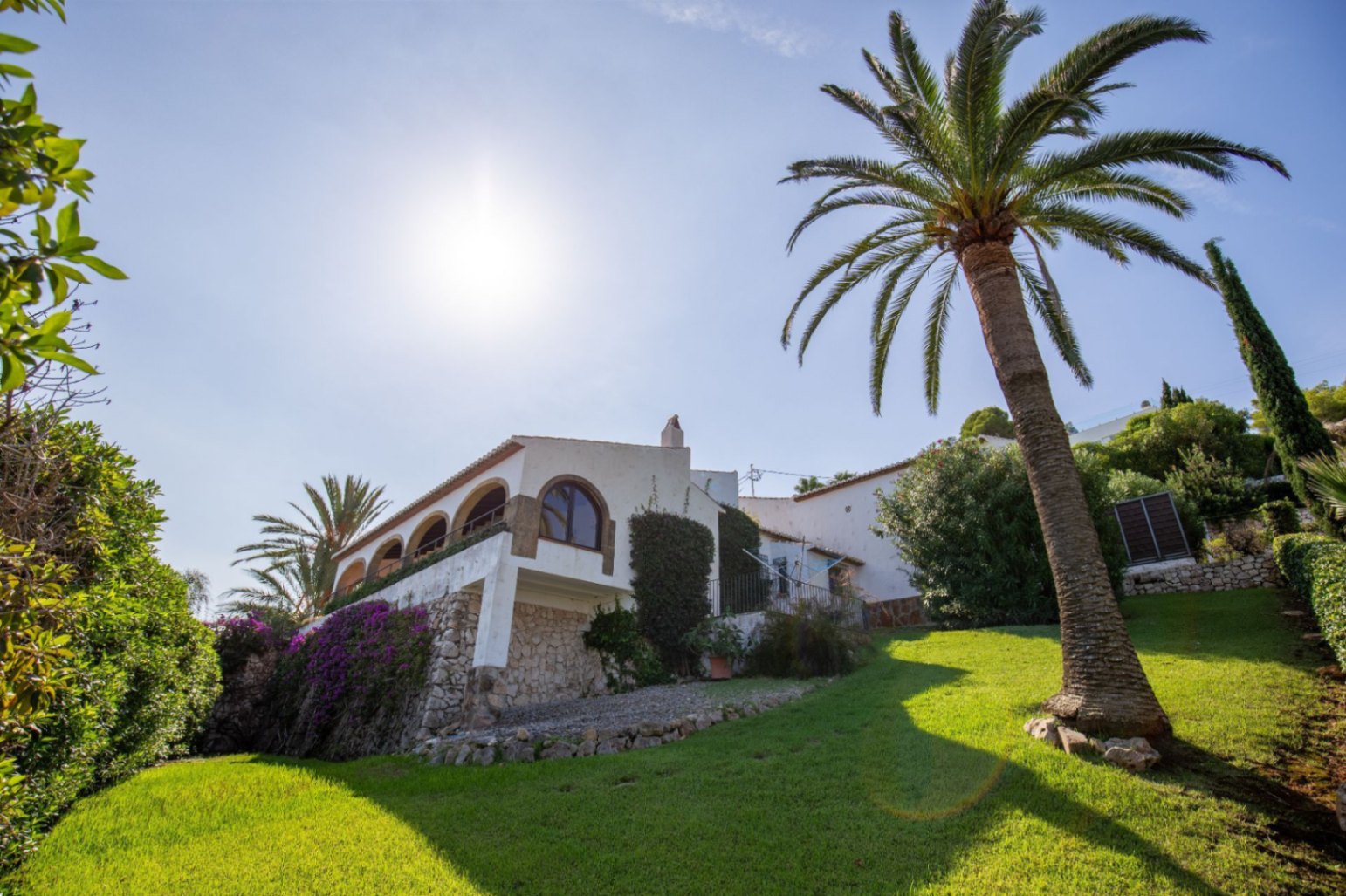 Außenansicht einer mediterranen Villa mit Rundbogenterrasse, Natursteinmauern und gepflegtem Rasenhang mit einer markanten Palme.