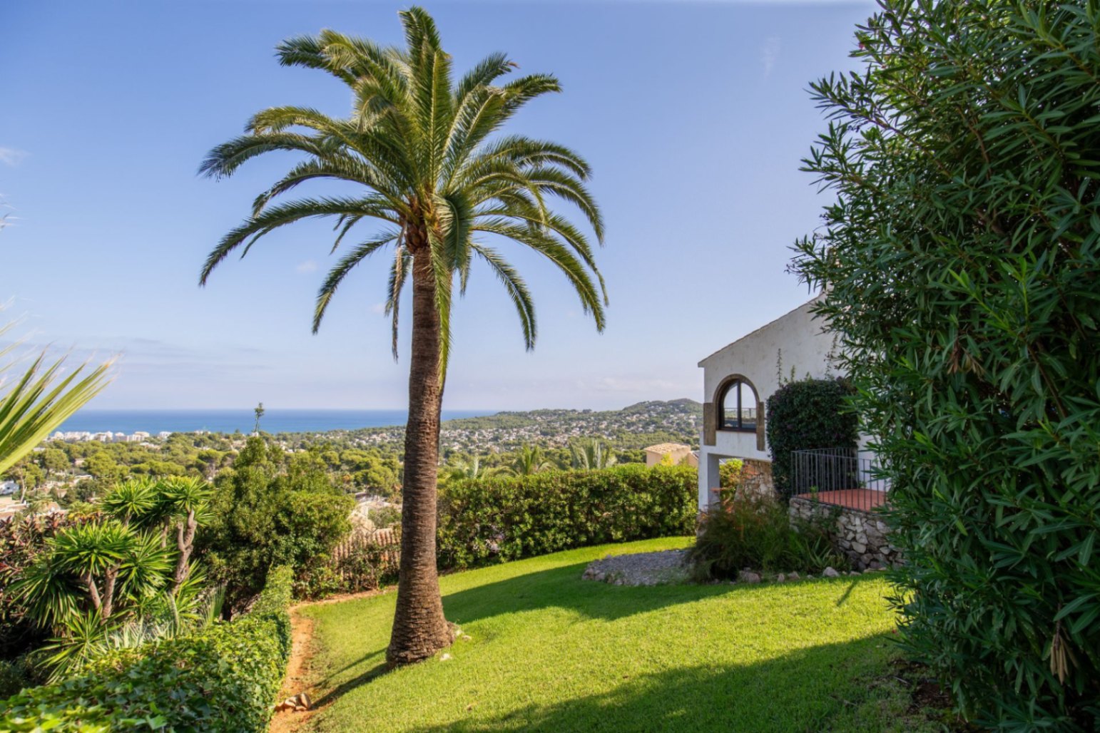 Garten mit einer ausgewachsenen Palme und Rasenfläche, Blick auf die Steinfassade und das Rundbogenfenster der Villa. Panorama-Küsten- und Meerblick im Hintergrund.