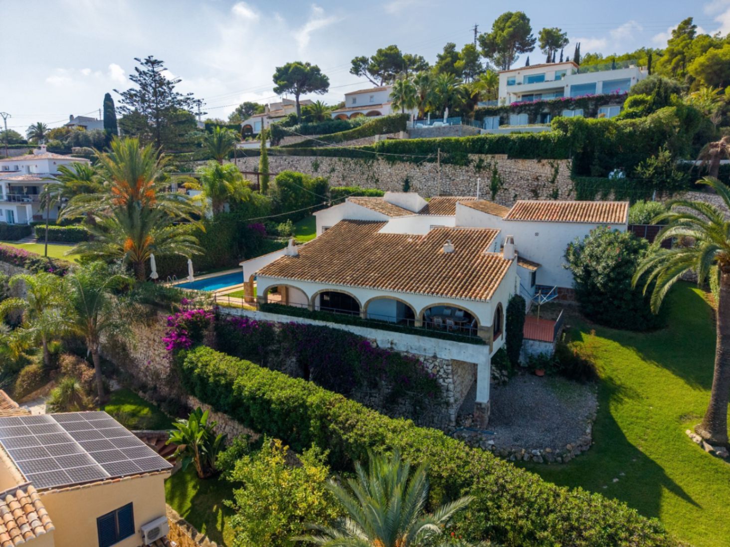 Luftaufnahme einer mediterranen Villa mit Terrakottadach, Bogenterrasse und Pool, eingebettet in terrassierte Steinmauern und Gärten mit Palmen.