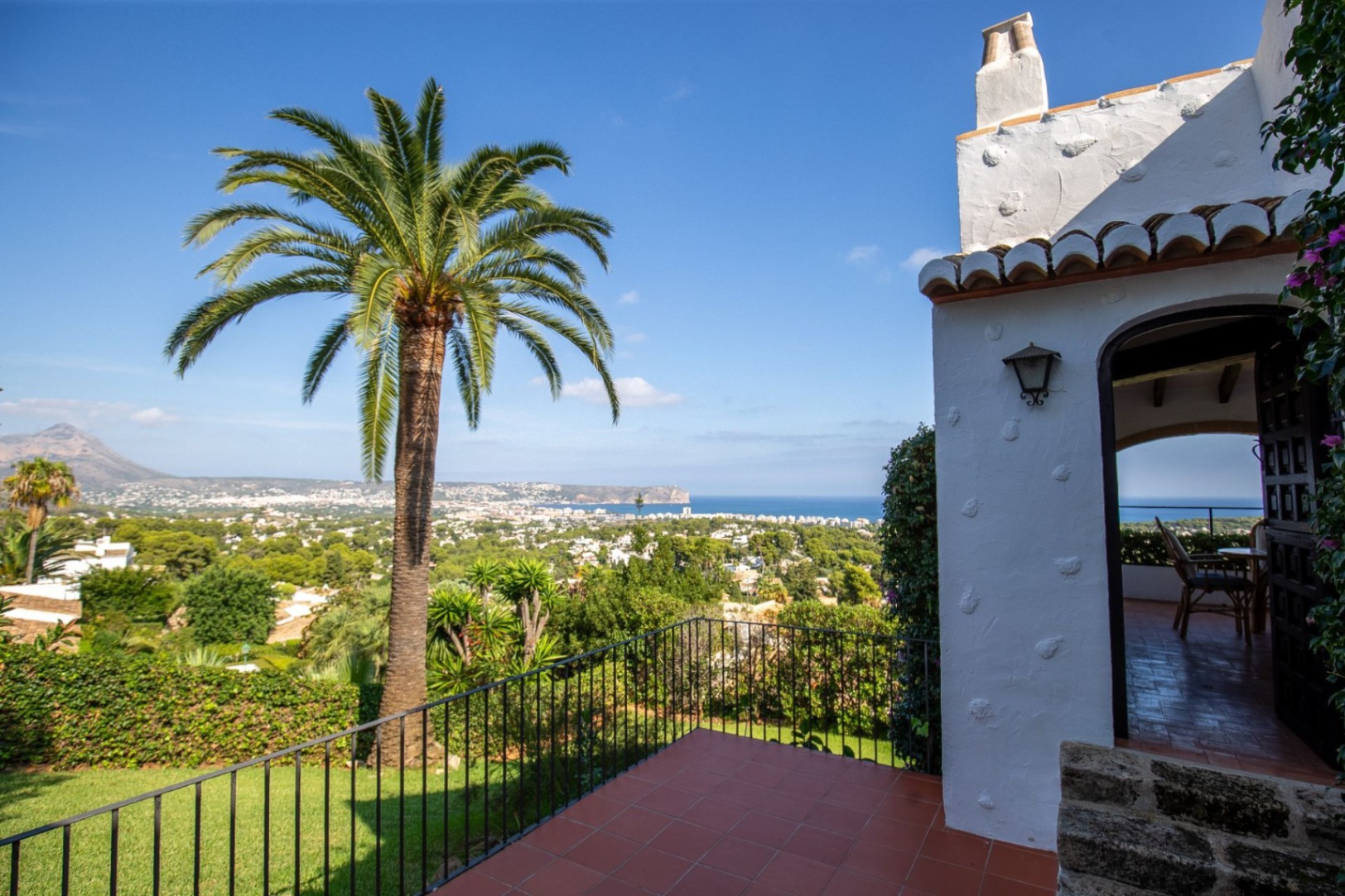 Terrasse mit Terrakotta-Fliesen und Panoramablick auf die Mittelmeerküste, akzentuiert durch eine große Palme und traditionelle weiße Architektur.