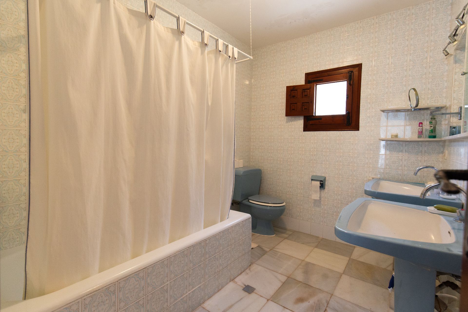 Traditionelles Badezimmer mit zwei blauen Säulenwaschbecken, passender Toilette und gefliester Badewanne. Mit gemusterten Wandfliesen und einem kleinen Holzfenster.