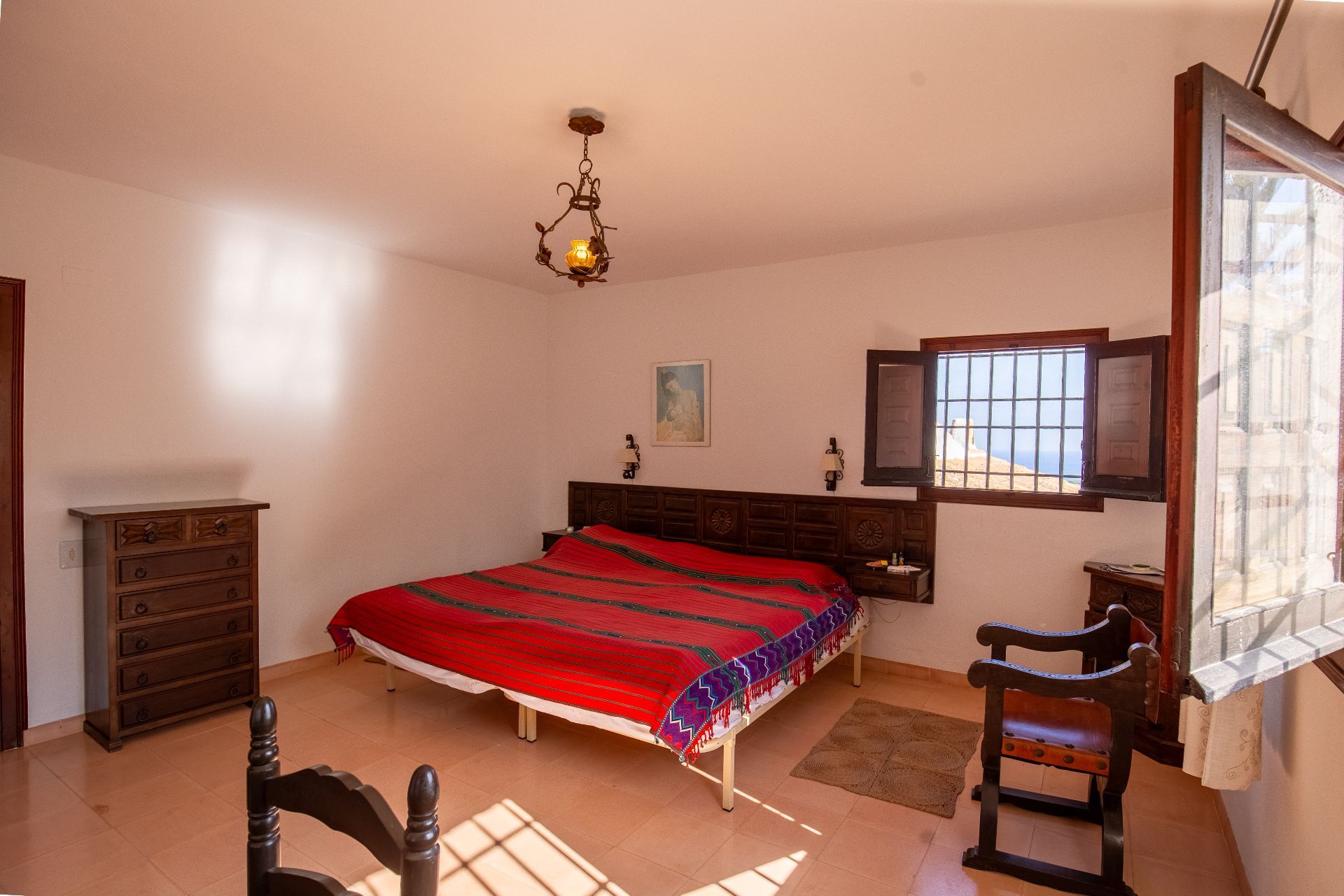 Traditionelles Schlafzimmer mit geschnitztem Holzkopfteil, Terrakottaboden und rustikalem Schmiedeeisen-Kronleuchter. Ein Fenster mit Holzläden sorgt für natürliches Licht.