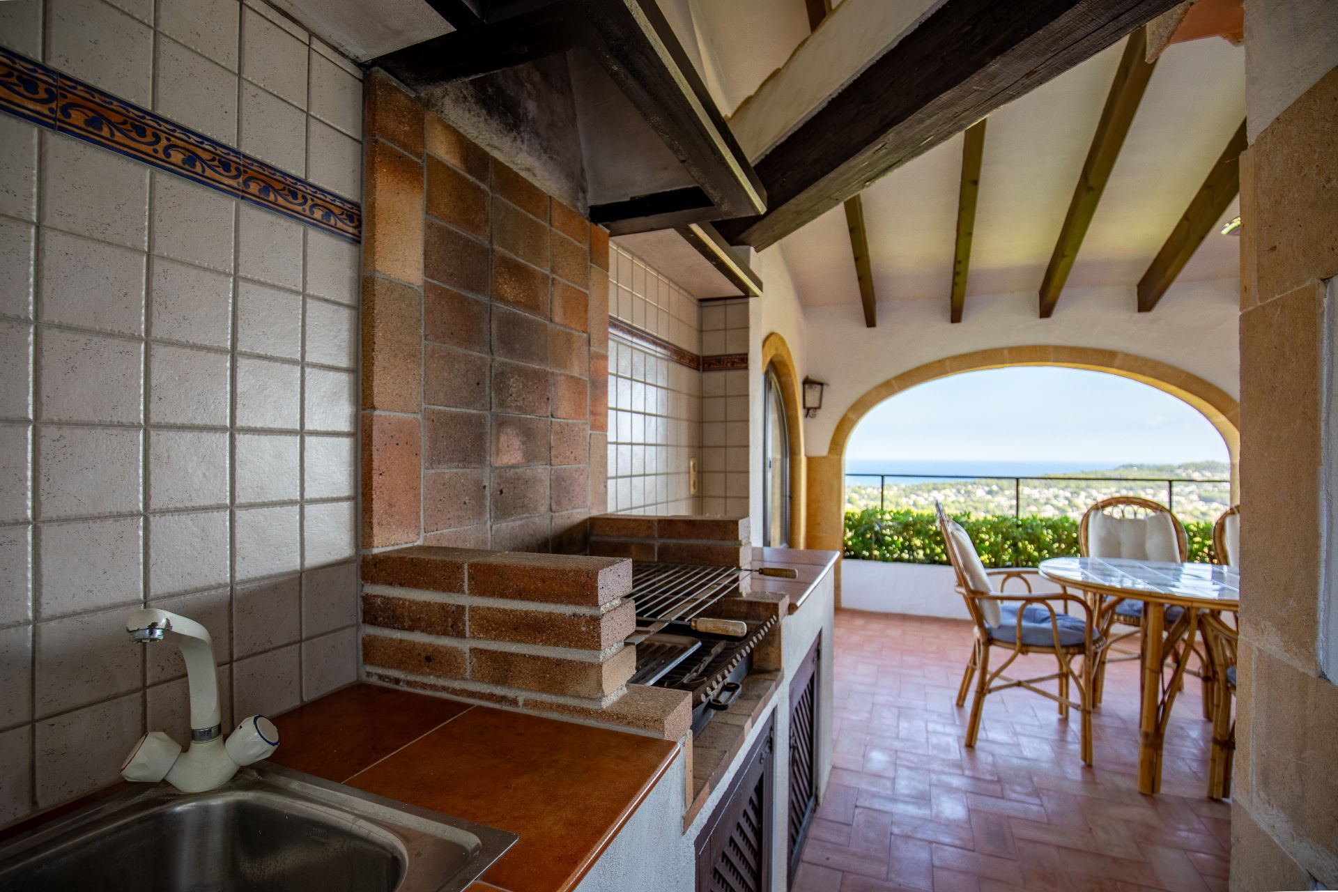 Überdachte Außenküche mit gemauertem Grill, Spülbecken und Terrakottaboden, ergänzt durch Holzbalkendecken und einen weiten Meerblick.