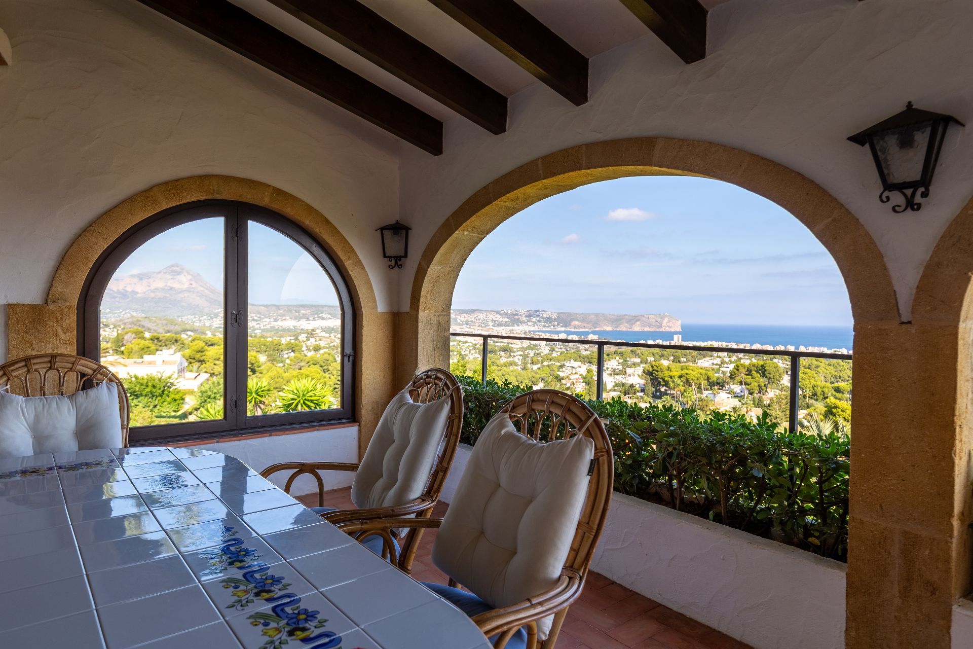 Überdachte Terrasse mit Steinbögen, Holzbalken und einem gefliesten Esstisch. Von den Korbsesseln aus bietet sich ein Panoramablick auf das Mittelmeer und die Berge.