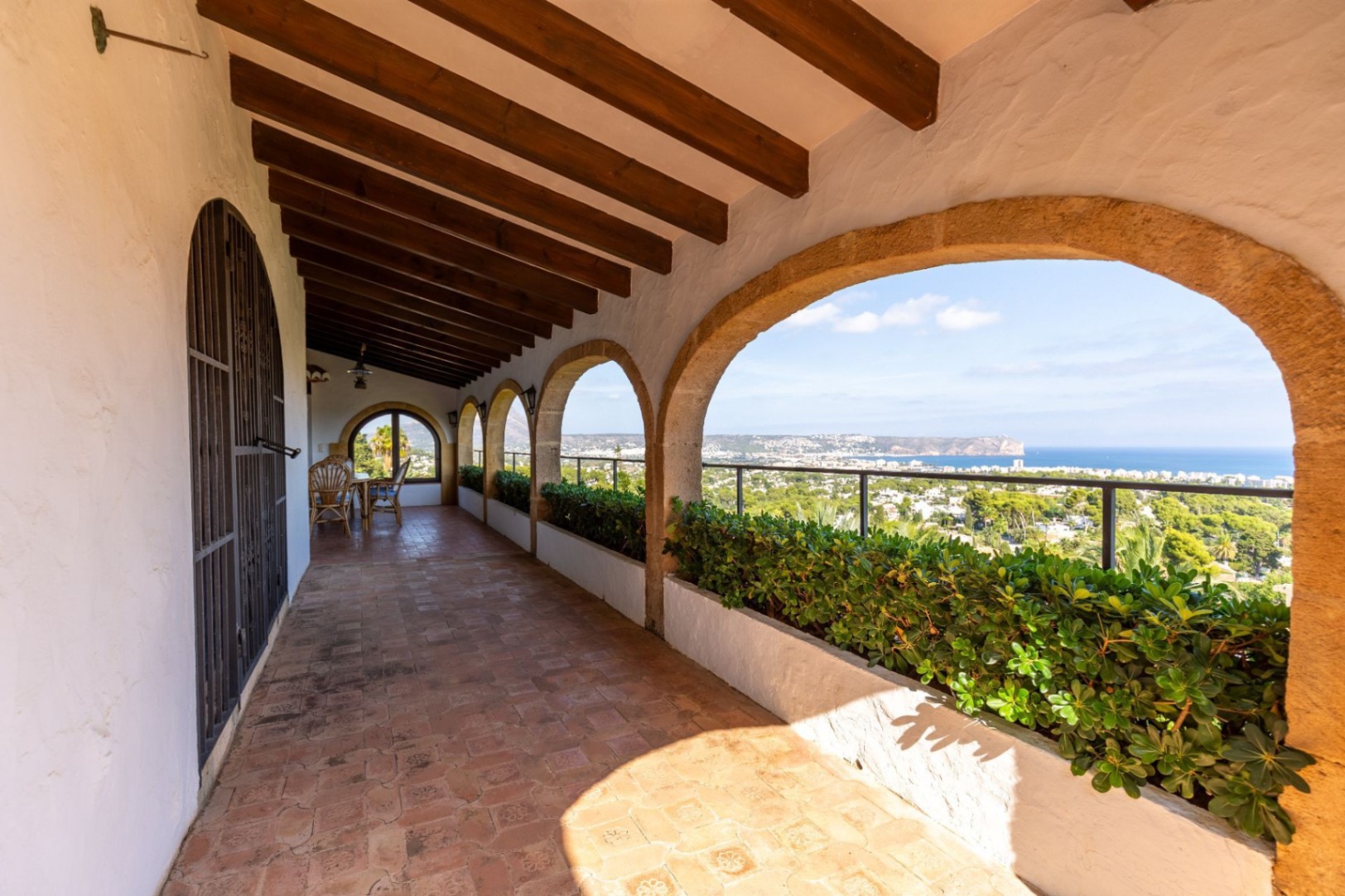 Überdachte Terrasse mit Steinbögen, sichtbaren Holzbalken und Terrakottaboden. Integrierte Pflanzgefäße rahmen den Panoramablick auf die Küste und das Meer ein.