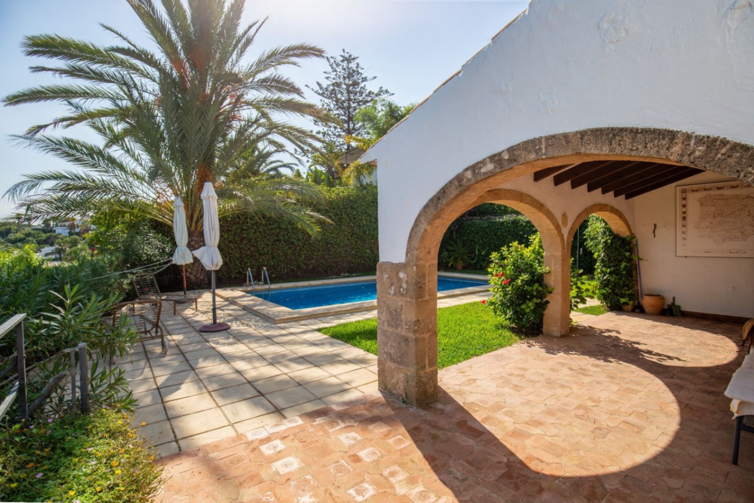 Überdachte Terrasse mit Steinbögen und Terrakottaboden mit Blick auf einen privaten Pool, umgeben von Palmen und mediterraner Vegetation.