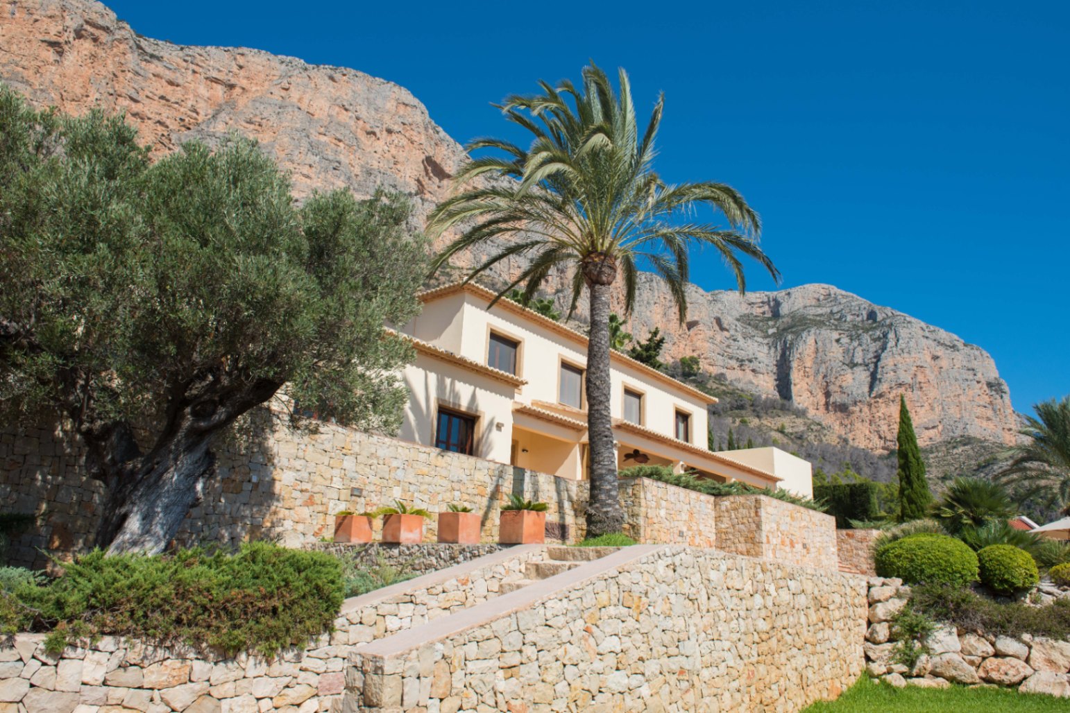 Außenansicht einer mediterranen Villa mit Natursteinmauern, einem Olivenbaum und einer Palme vor der Kulisse einer markanten Felsformation.