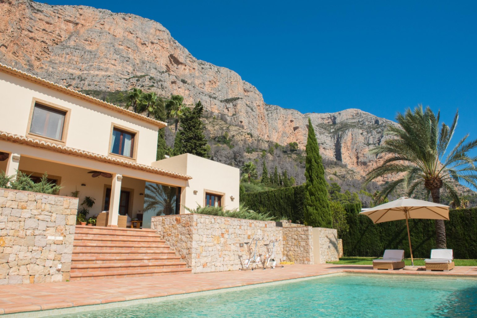 Außenbereich mit türkisfarbenem Pool, Terrakotta-Terrasse und Natursteinmauern vor einer markanten Bergkulisse mit Palmen und Zypressen.