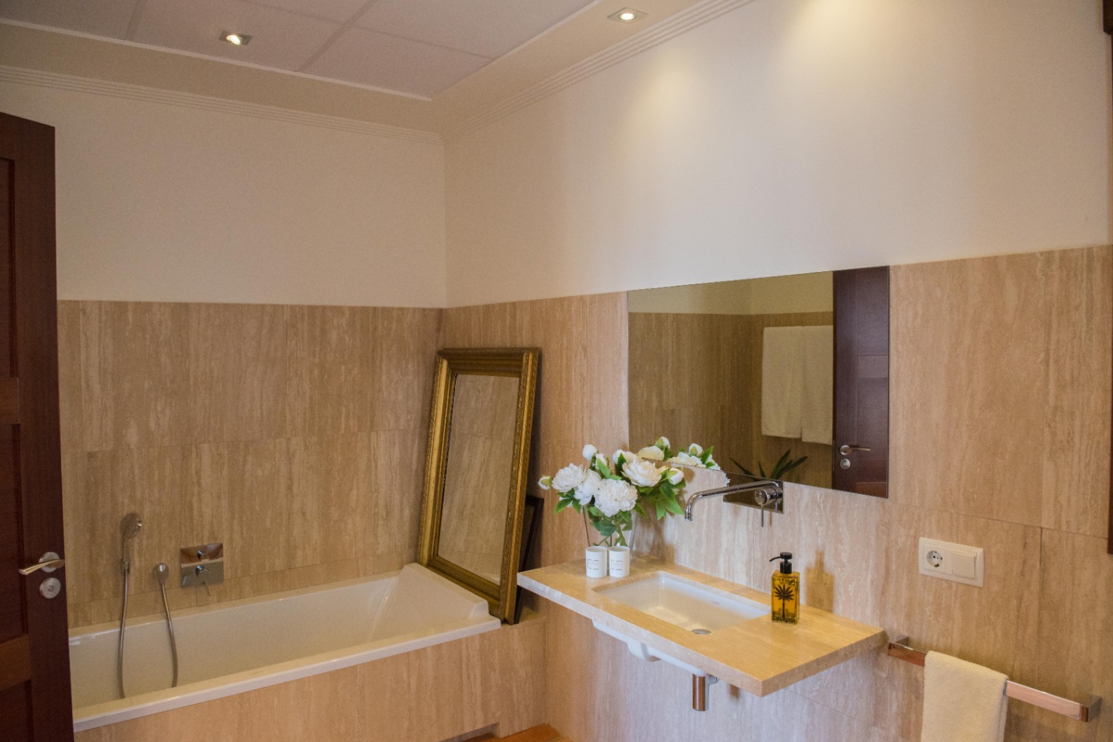 Badezimmer mit Travertinwänden, schwebendem Steinwaschtisch und Einbaubadewanne. Akzentuiert durch einen goldgerahmten Spiegel und Deckeneinbaustrahler.