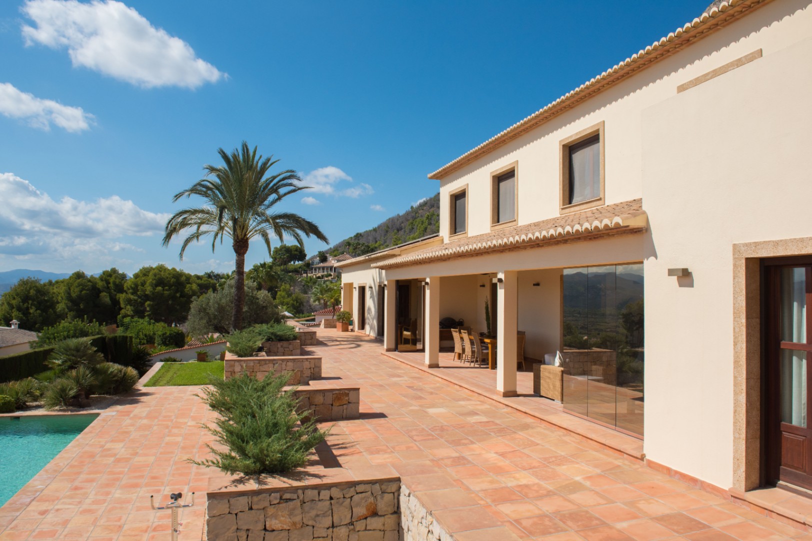 Mediterrane Villa mit Terrakotta-Terrasse, Naturstein-Mauern und überdachter Veranda. Eine Palme ziert den Außenbereich neben dem Pool mit Blick auf die Berge.