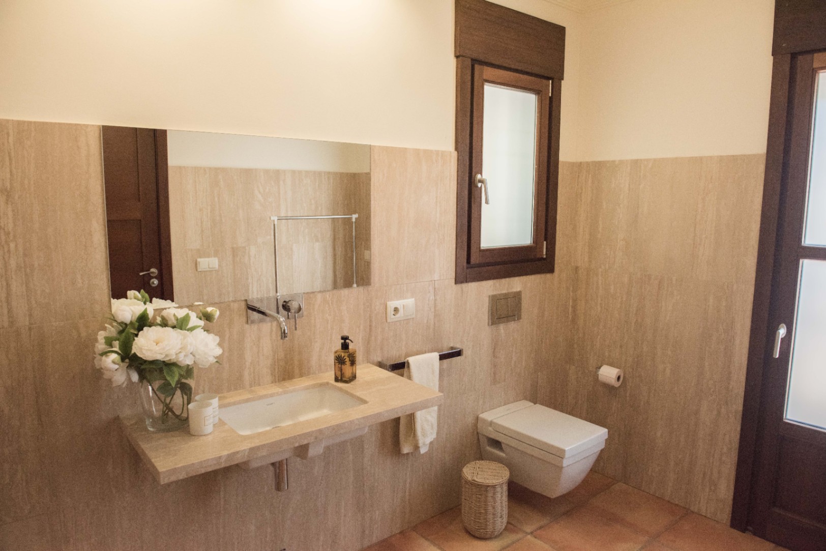 Modernes Badezimmer mit schwebendem Steinwaschtisch, wandmontierten Armaturen und wandhängendem WC vor hellen Travertinwänden und Terrakottaboden.