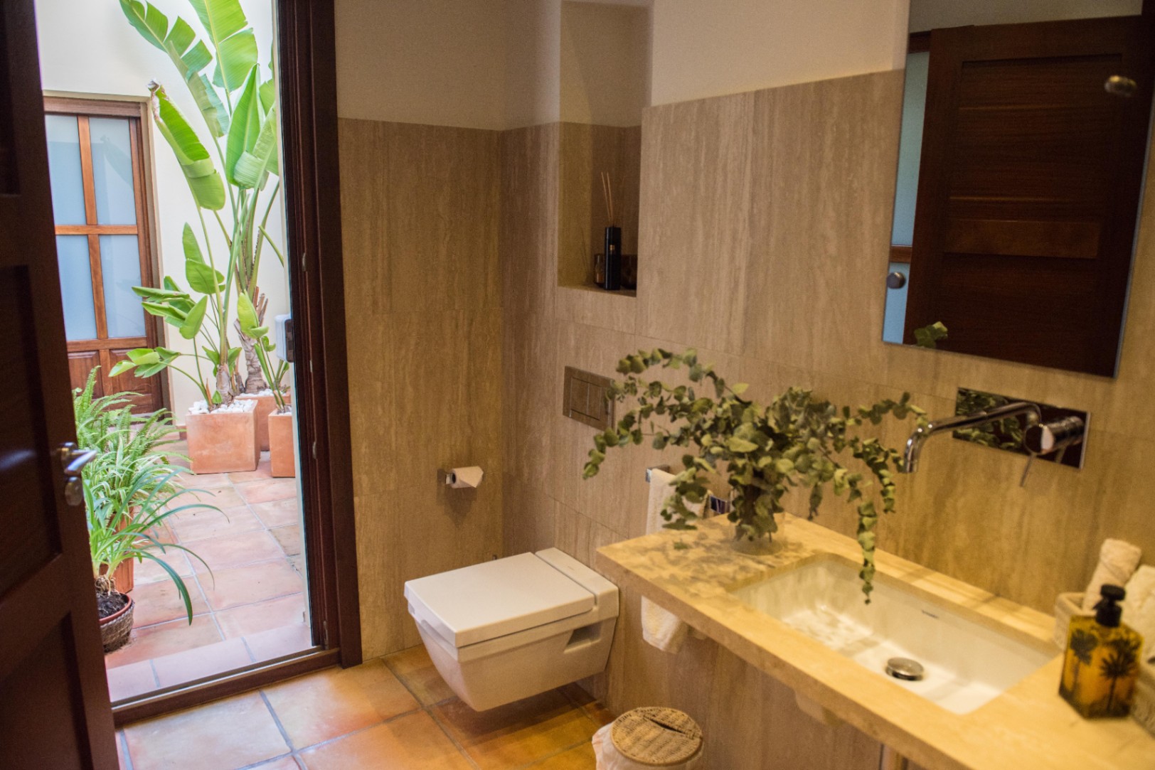 Modernes Badezimmer mit Travertin-Wänden, wandhängendem WC und Zugang zu einem privaten Innenhof mit Terrakotta-Fliesen und üppiger Bepflanzung.