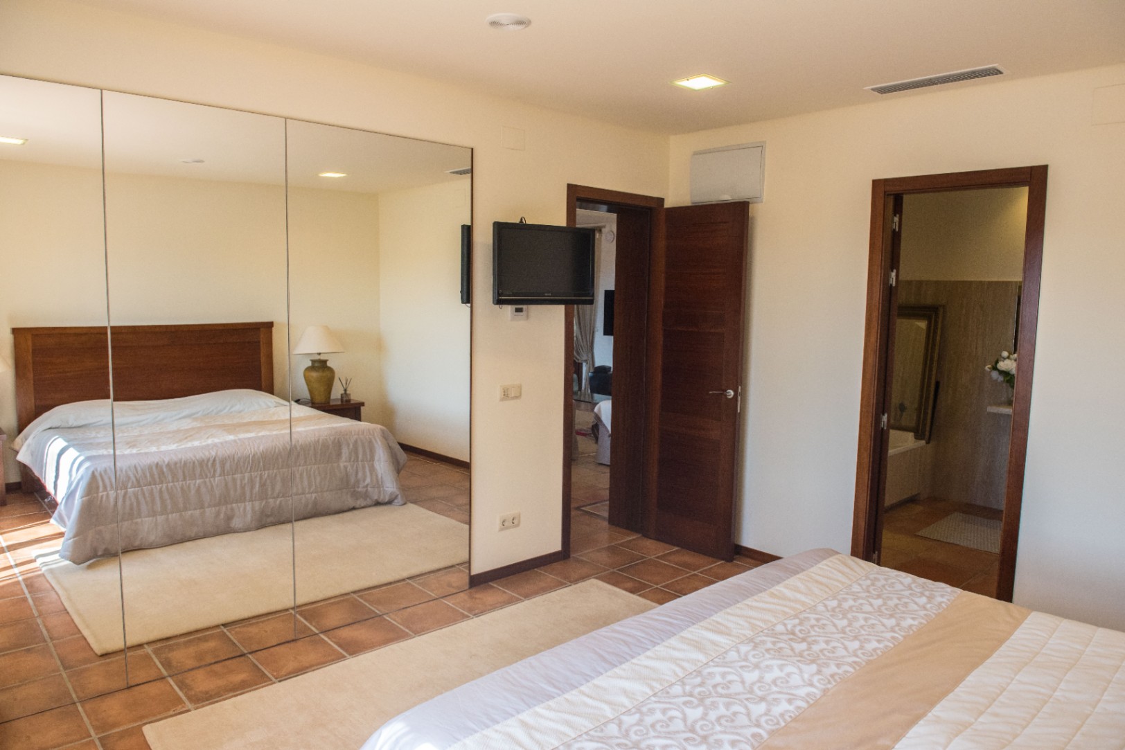 Schlafzimmer mit Terrakotta-Fliesen, großem Spiegelschrank und Wand-TV. Der Raum verfügt über Deckeneinbaustrahler und einen direkten Zugang zum angrenzenden Badezimmer.