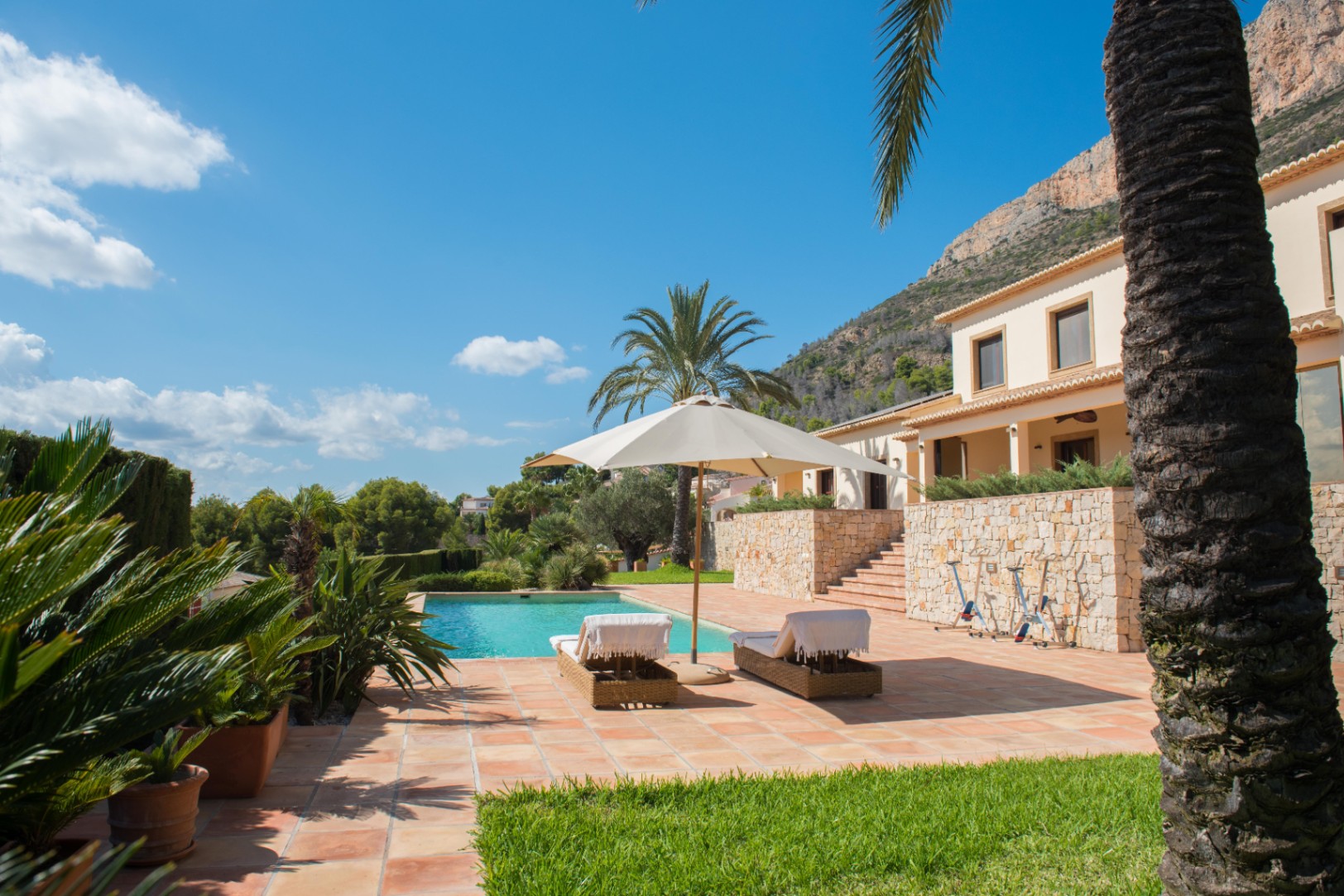 Terracotta-Terrasse mit rechteckigem Pool, Korbliegen und Natursteinmauern. Die mediterrane Gartenanlage bietet Palmen und einen direkten Blick auf das angrenzende Bergmassiv.